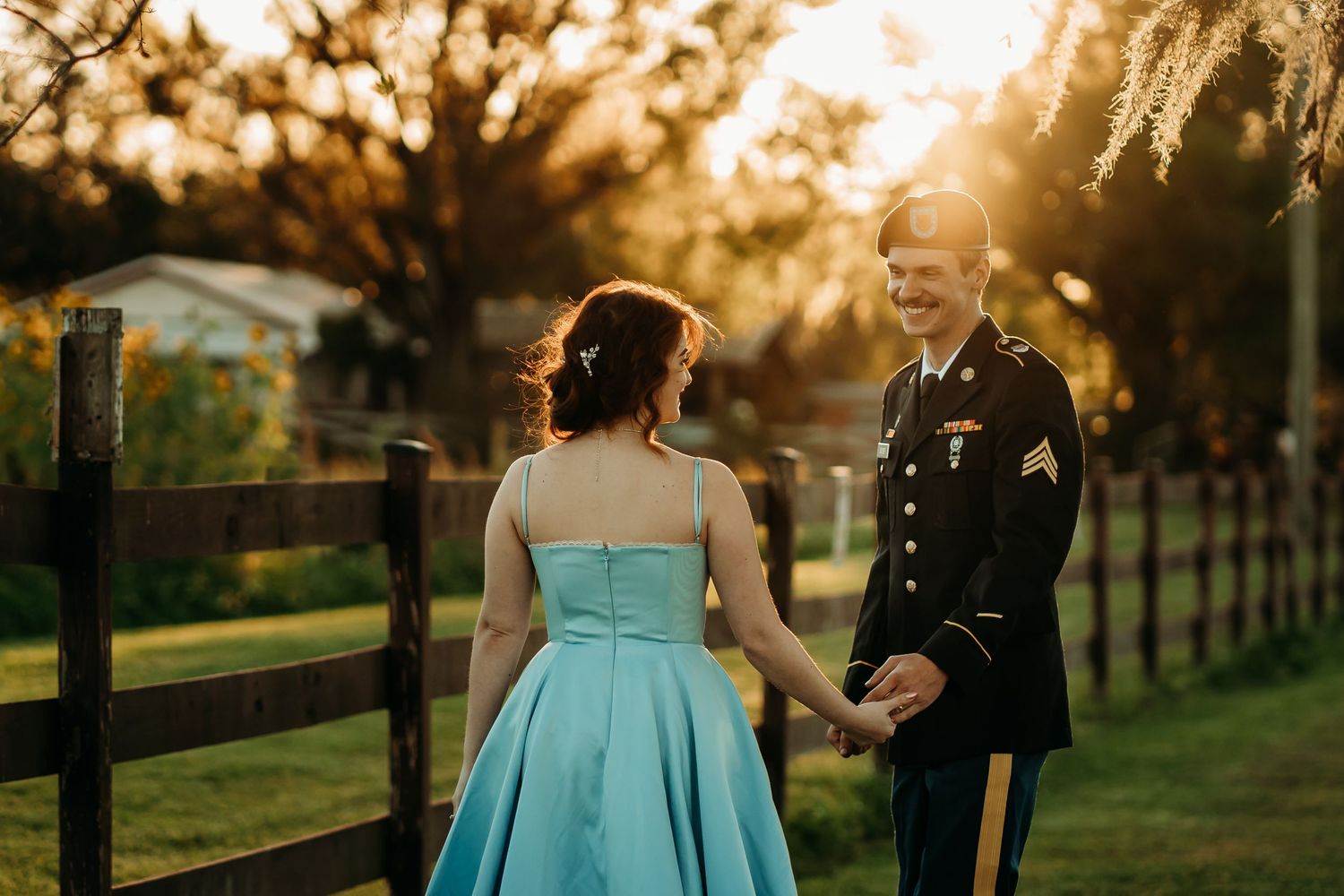 Soldier in Army dress uniform and woman in blue gown walk hand in hand at golden hour.