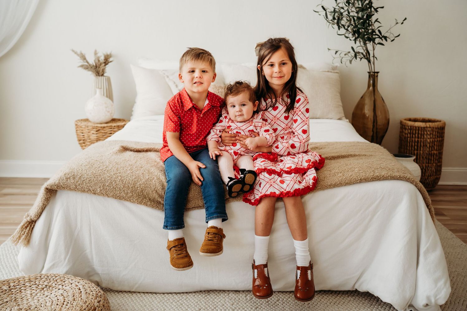 Three young children sitting together on a bed, dressed in red holiday outfits, smiling for a portrait.
