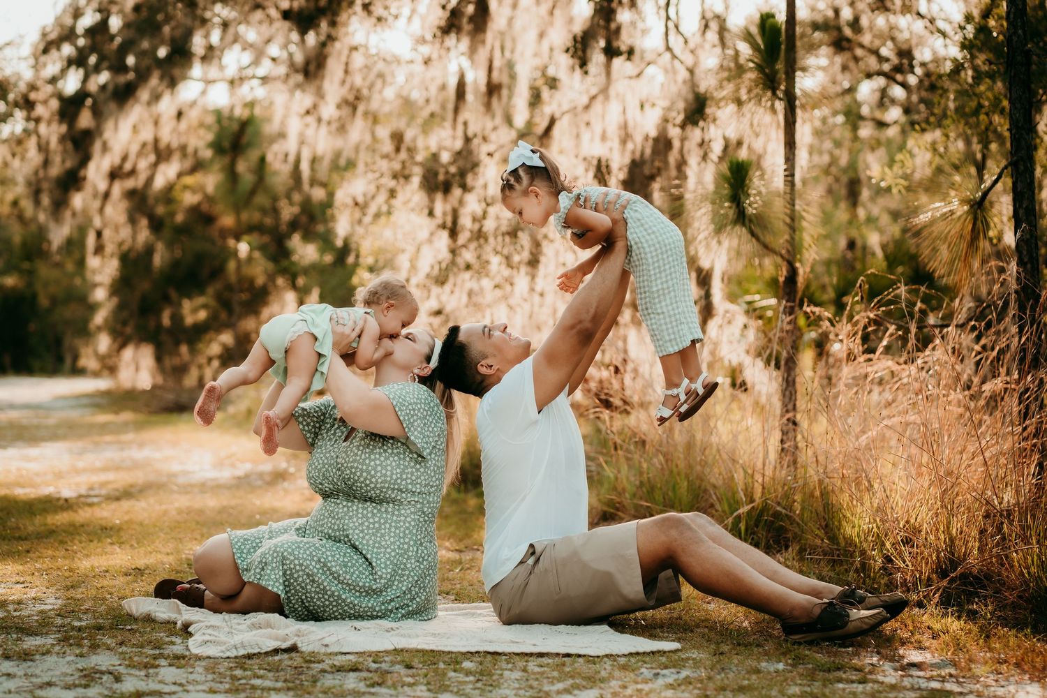 Parents lifting twin babies outdoors under Spanish moss-draped trees.