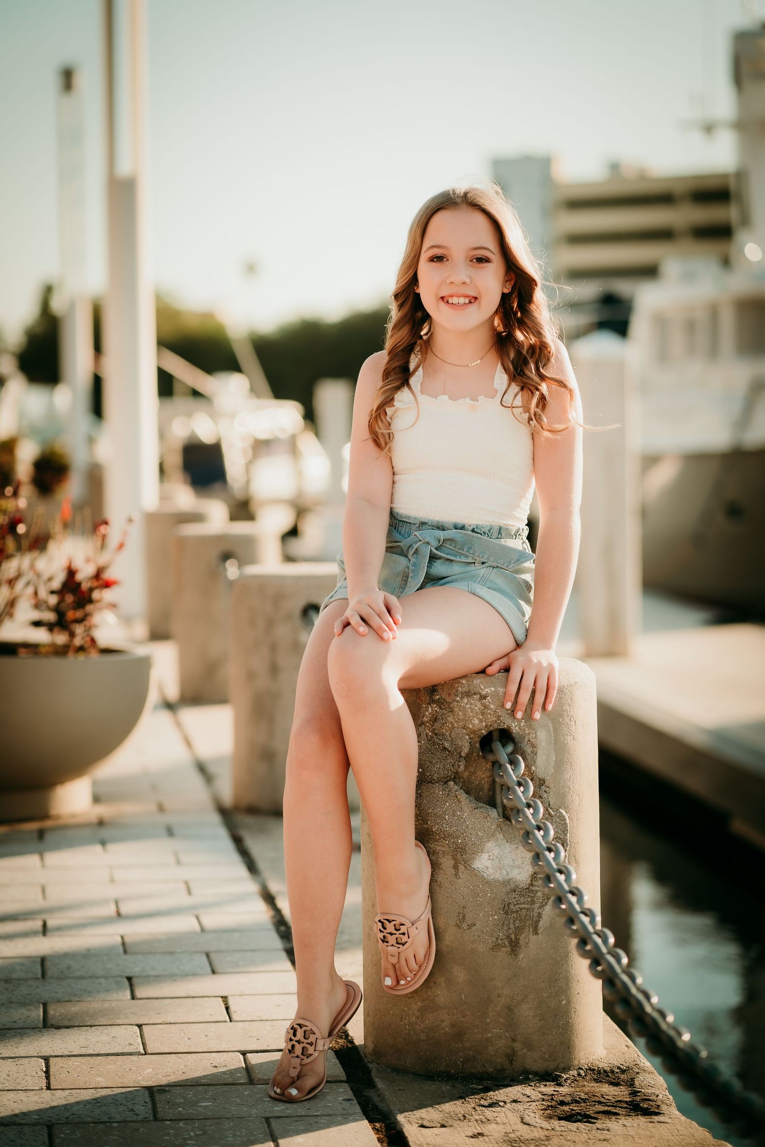 Young girl in white top and denim shorts sitting on a marina bollard, smiling in golden hour light.