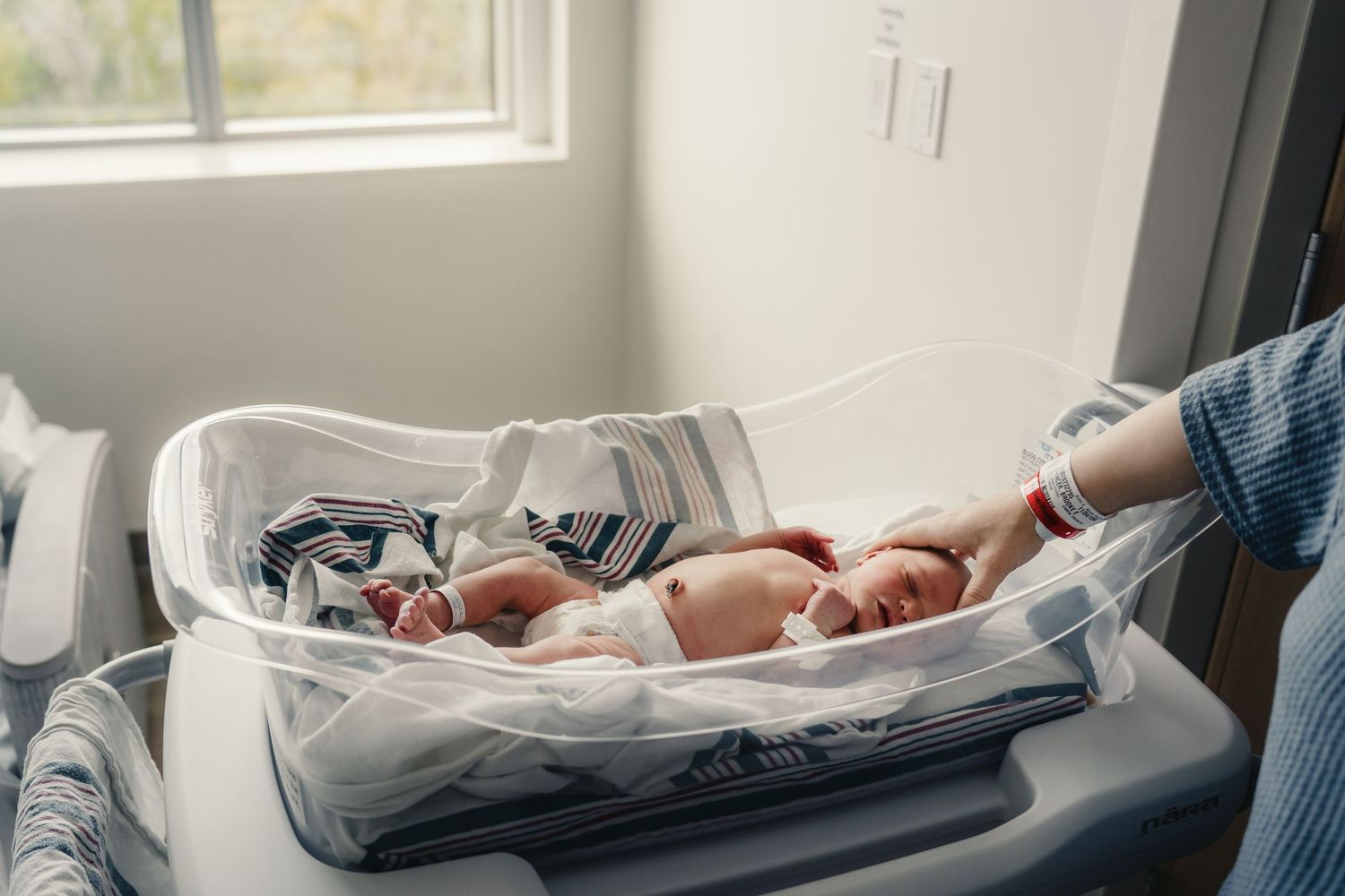A newborn baby rests in a clear hospital bassinet as a parent gently reaches in to touch them near a window.