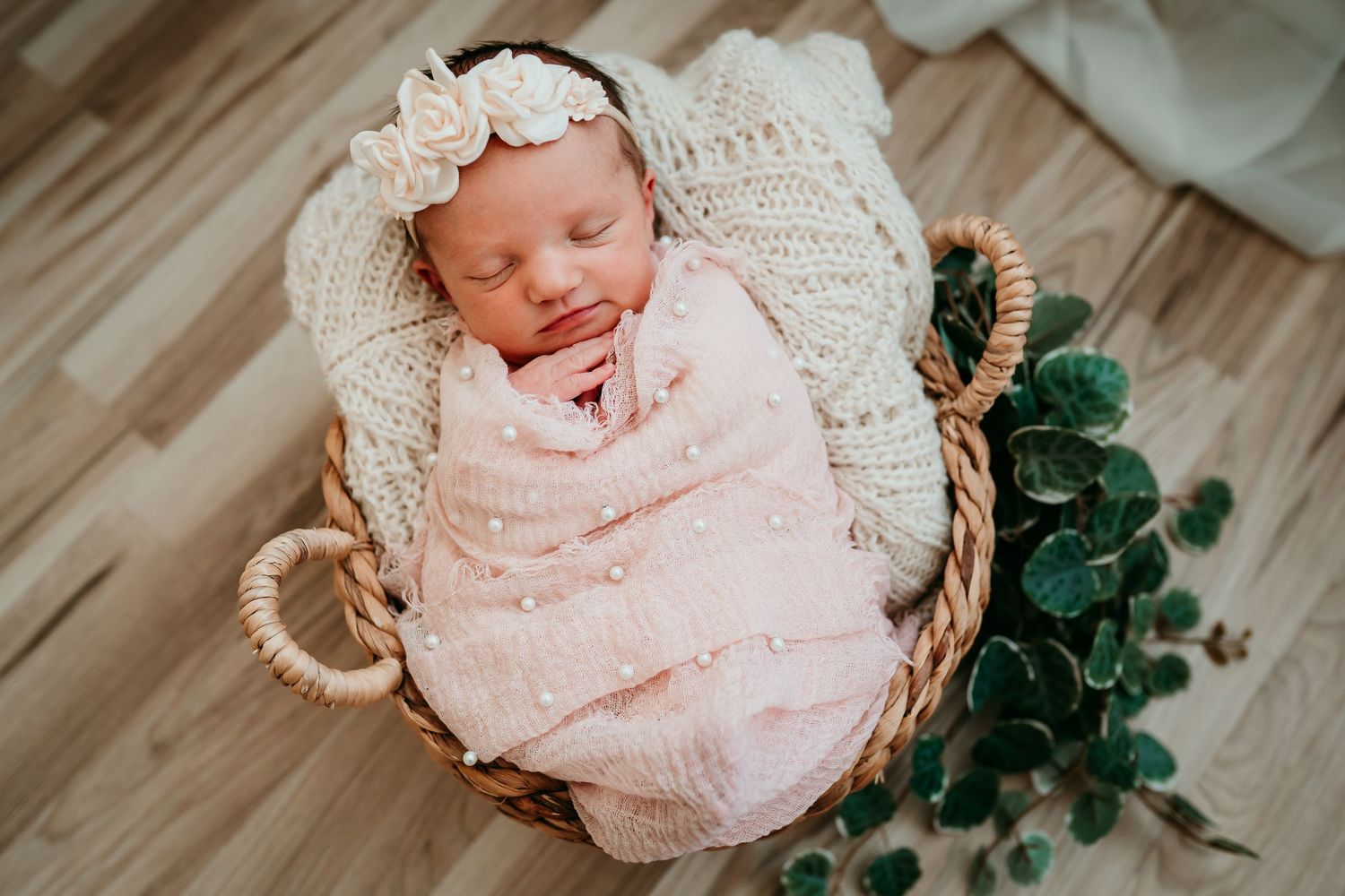 Sleeping newborn baby girl swaddled in pink wrap with floral headband in a wicker basket.