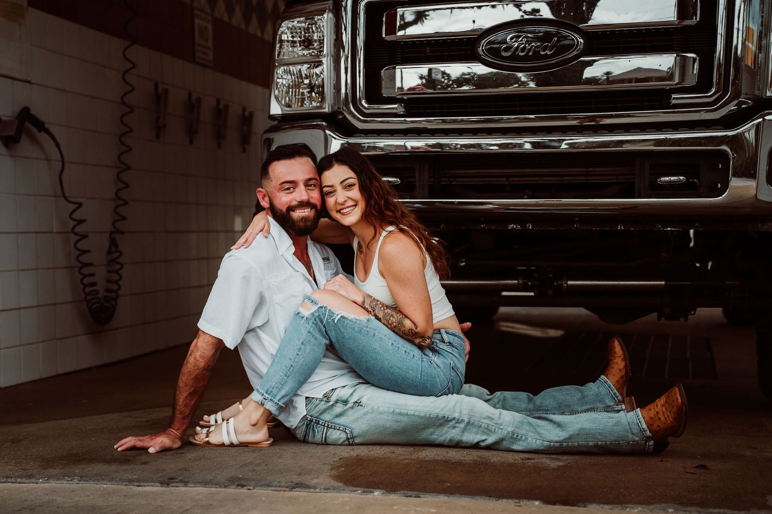 A couple sits together on a garage floor in front of a Ford truck, smiling warmly at the camera.