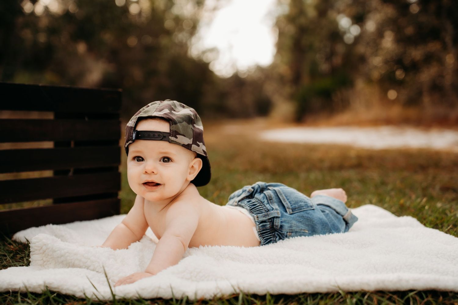 Adorable baby boy in camo hat and denim shorts lying on white blanket outdoors in a natural park setting.