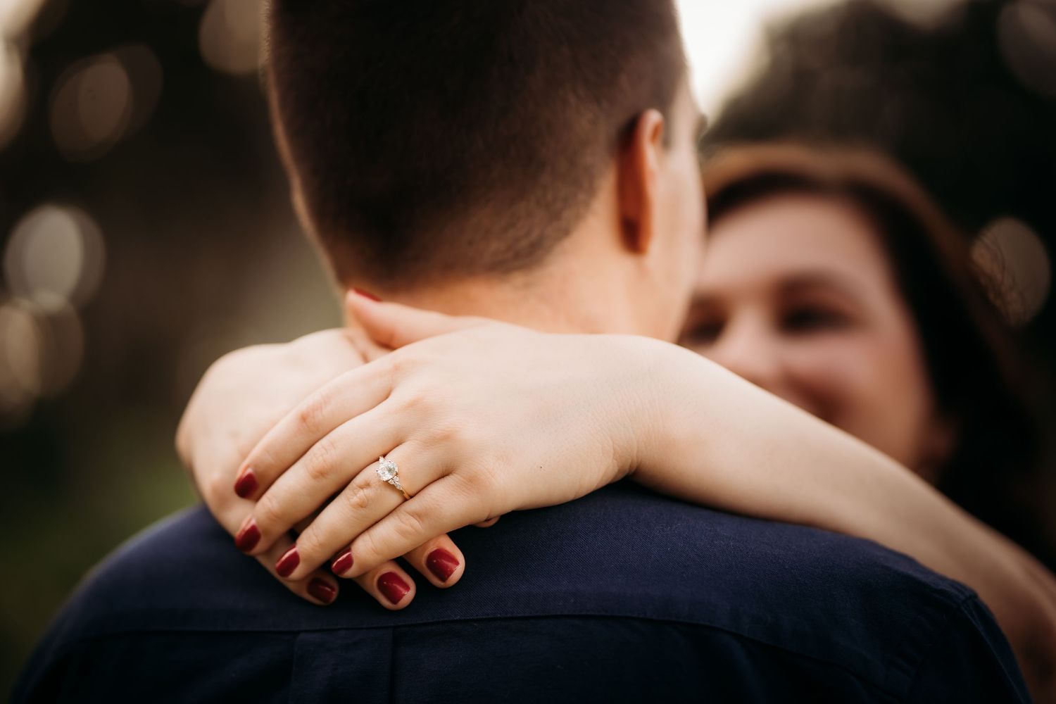 Close-up of a woman with red nails and an engagement ring embracing a man in a navy shirt outdoors.