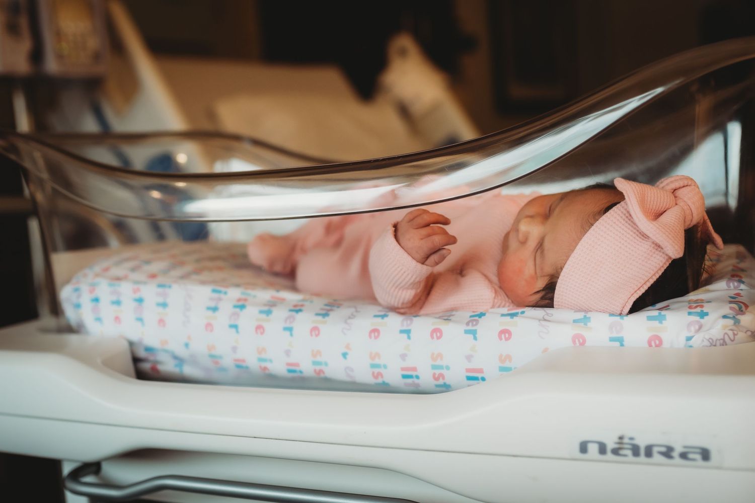 Newborn baby sleeping in Nara hospital bassinet wearing pink outfit and bow headband.