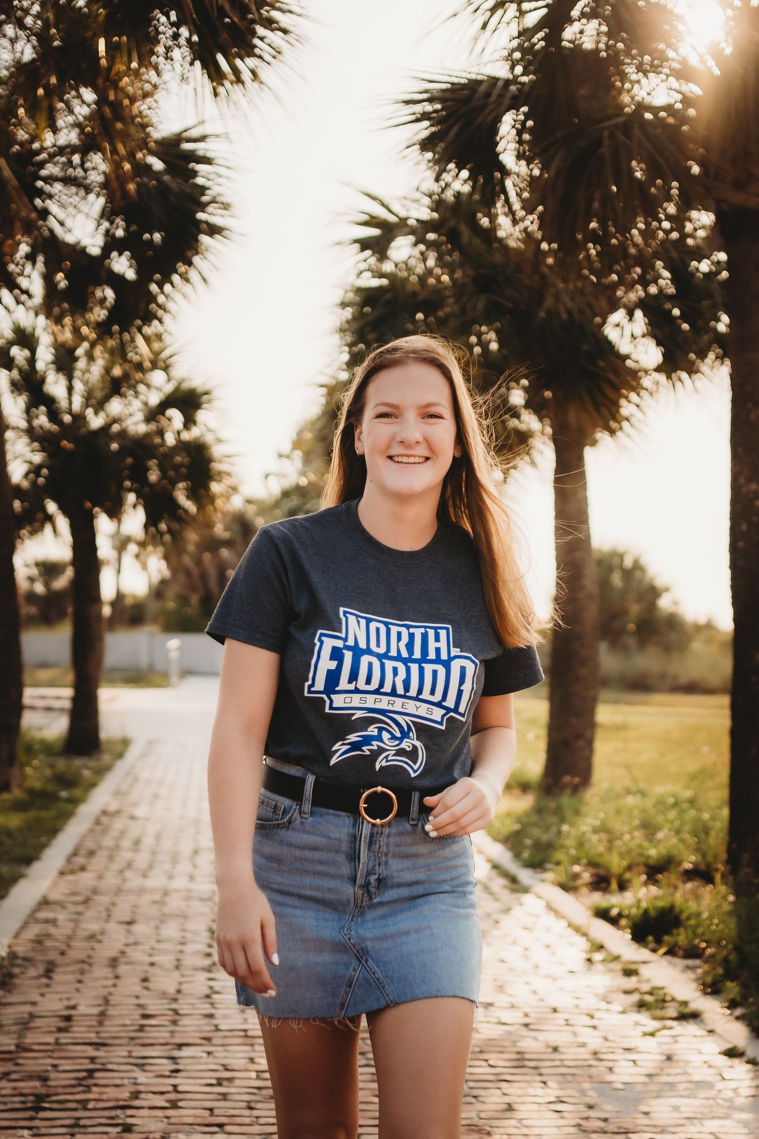 Smiling young woman wearing a University of North Florida t-shirt and denim skirt standing on a brick path lined with palm trees.