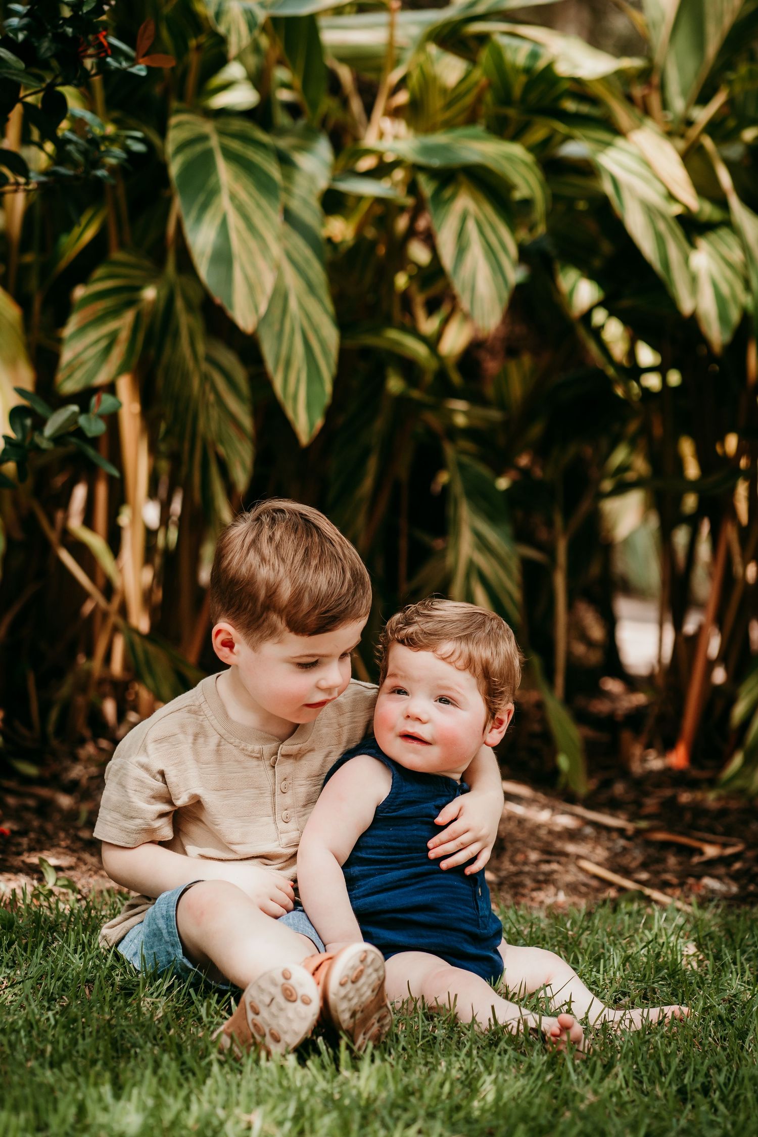 Older brother lovingly hugs baby sibling while sitting on grass near tropical plants.