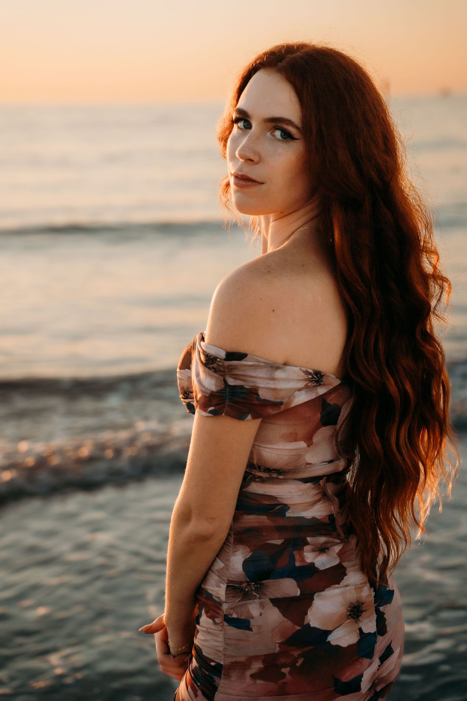 A woman with long red hair in a floral off-shoulder dress poses on a beach at sunset.