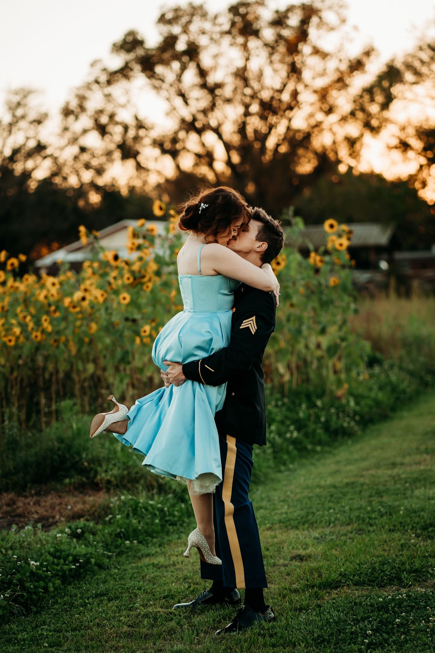 Soldier in dress uniform lifts woman in blue dress for a kiss near a sunflower field at sunset.