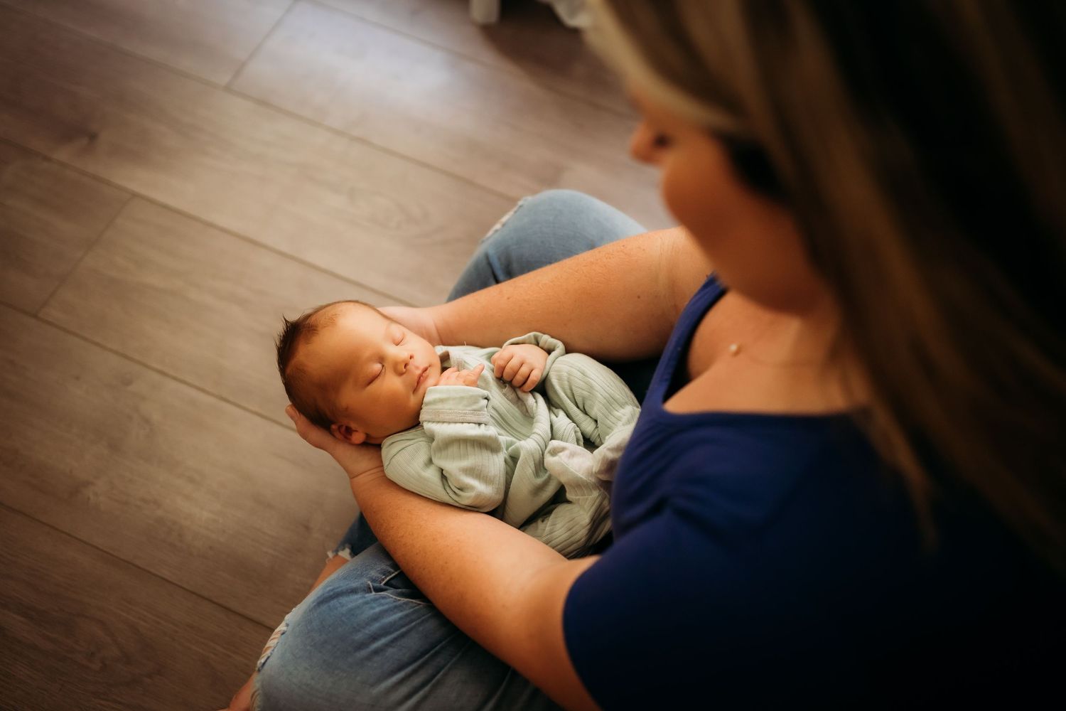 Mother sitting on floor cradling sleeping newborn baby dressed in soft gray outfit.