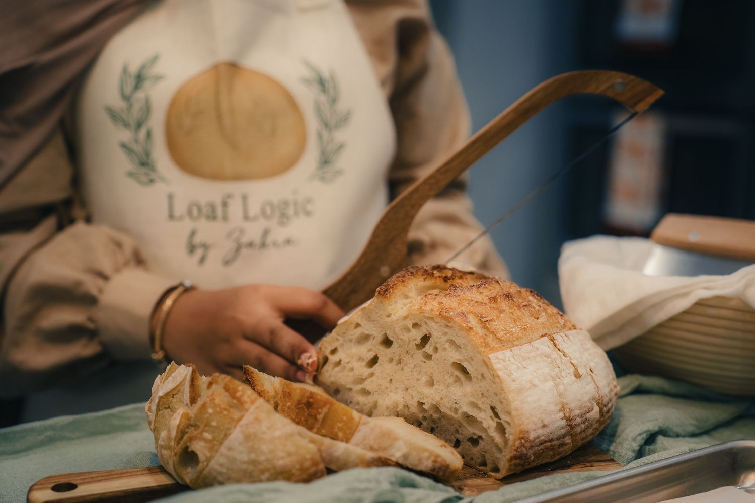 Person in Loaf Logic by Julia apron slicing fresh sourdough bread on a wooden cutting board.