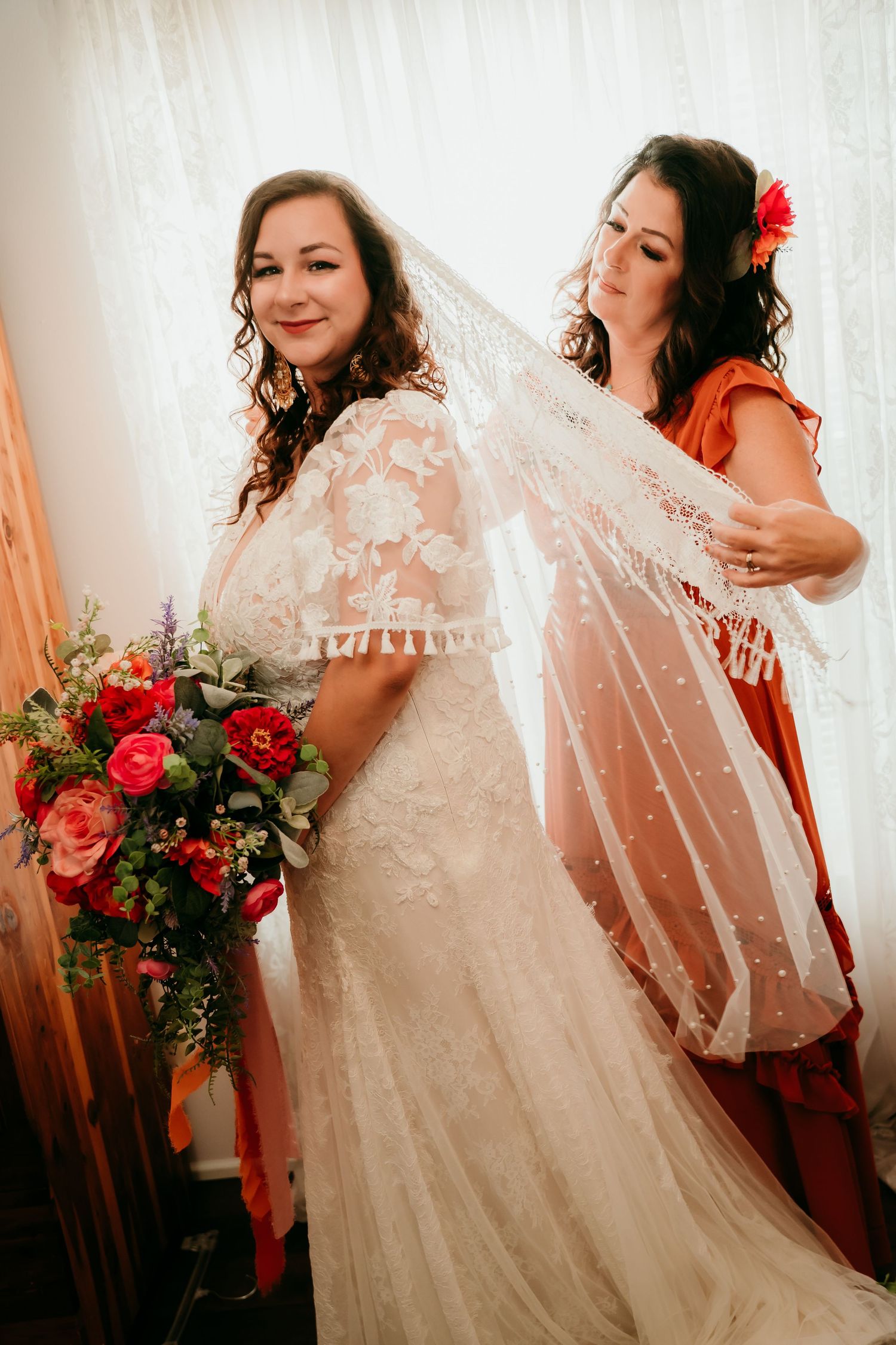 Bride in lace wedding gown holds red bouquet while bridesmaid in orange dress adjusts her polka dot veil.