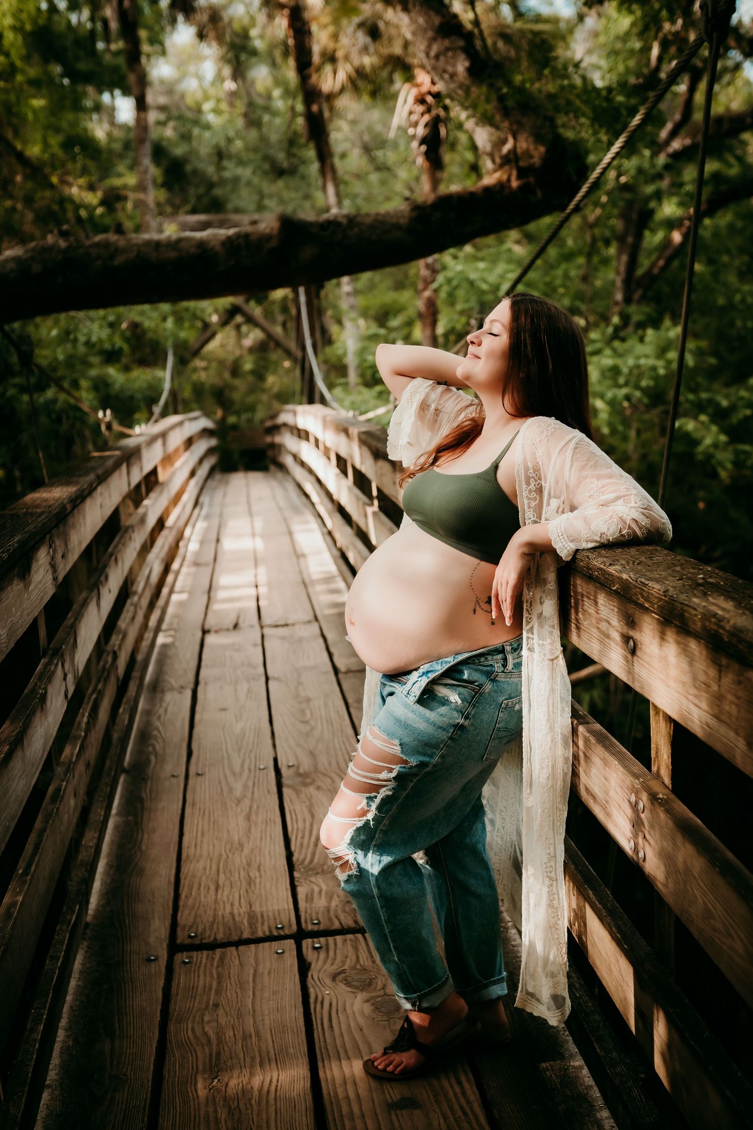 Pregnant woman in green bralette and ripped jeans posing on a rustic wooden bridge outdoors.