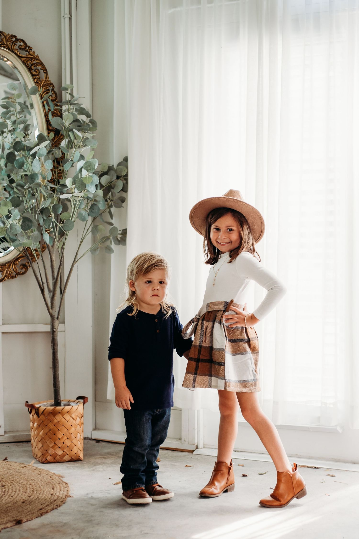 Two young children posing together indoors, girl in plaid skirt and hat standing confidently with younger boy.
