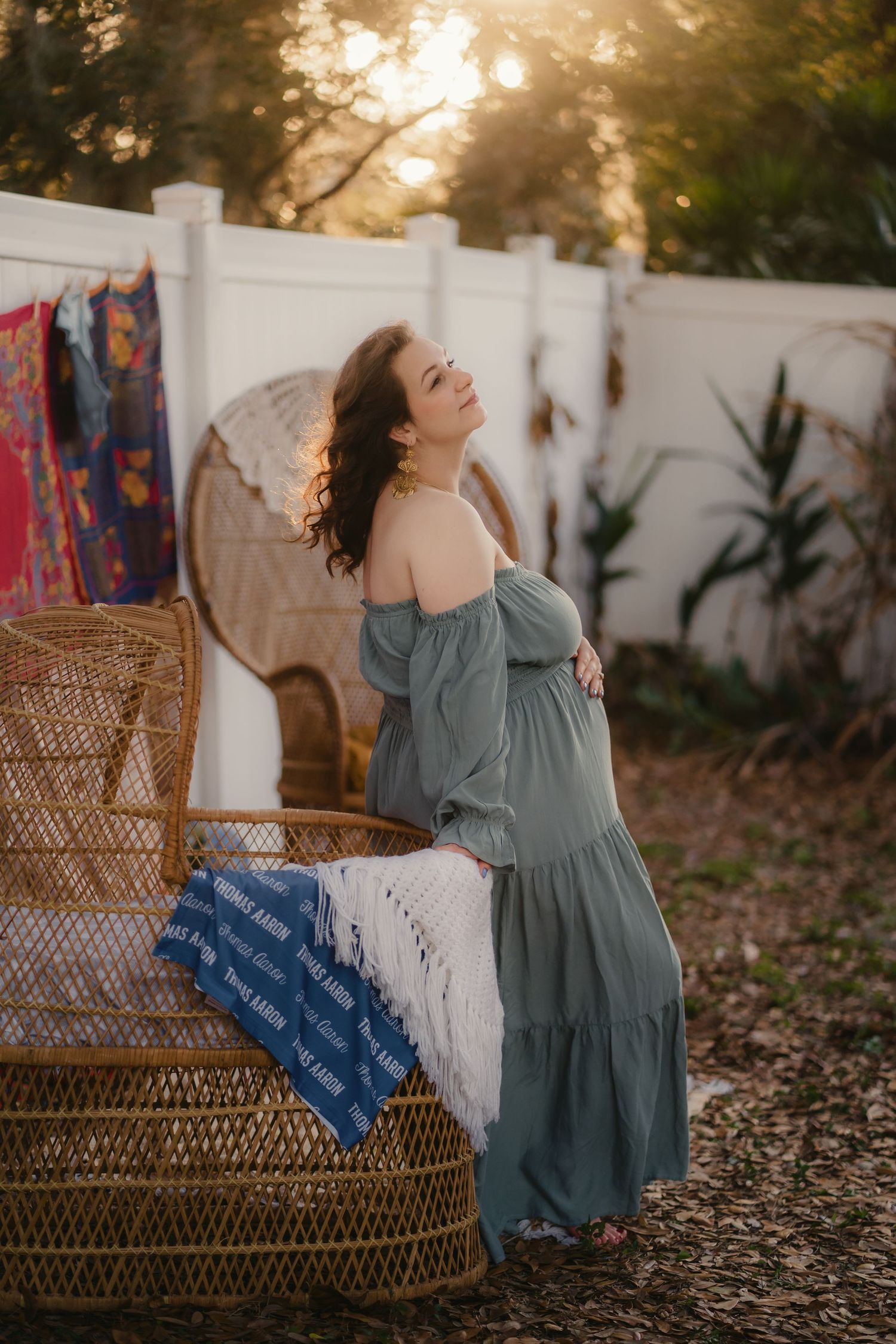 Pregnant woman in sage off-shoulder maxi dress poses by wicker peacock chair at golden hour in boho outdoor setting.