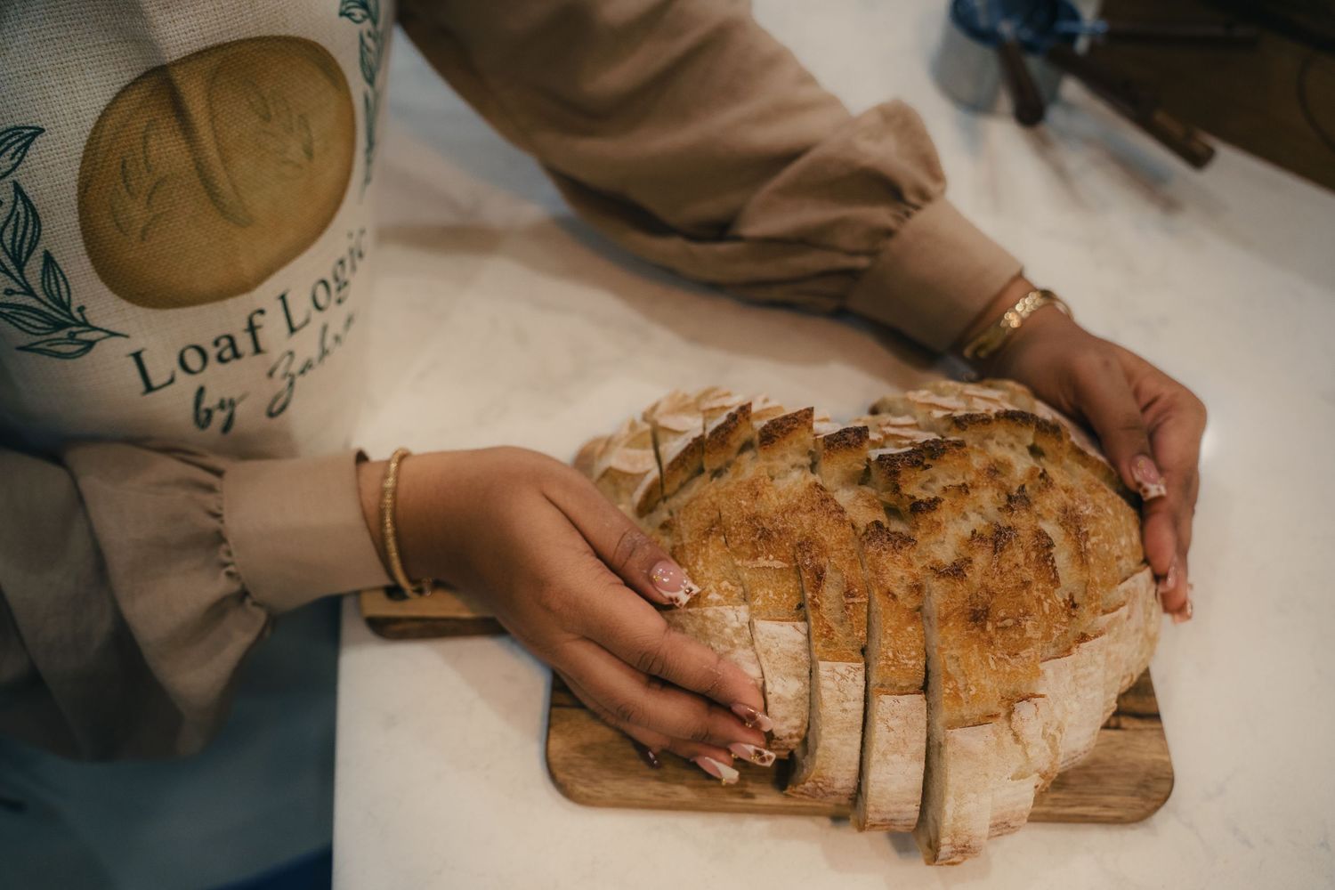 Woman slicing fresh sourdough bread on a wooden cutting board, wearing a Loaf Lounge apron and gold bracelets.