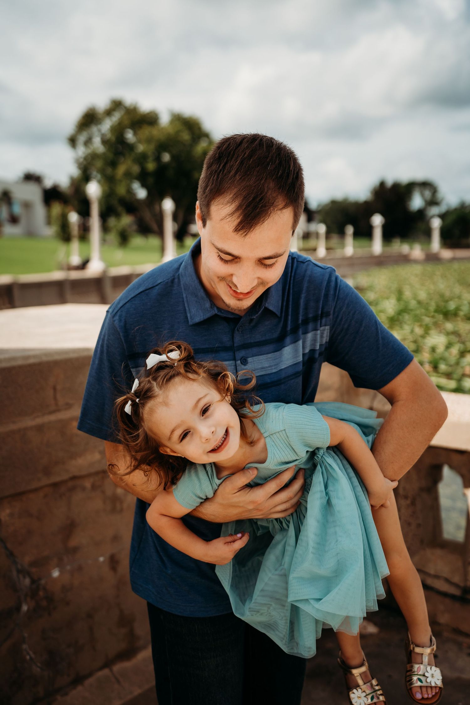 Smiling father playfully holding his laughing little girl in a teal dress outdoors.