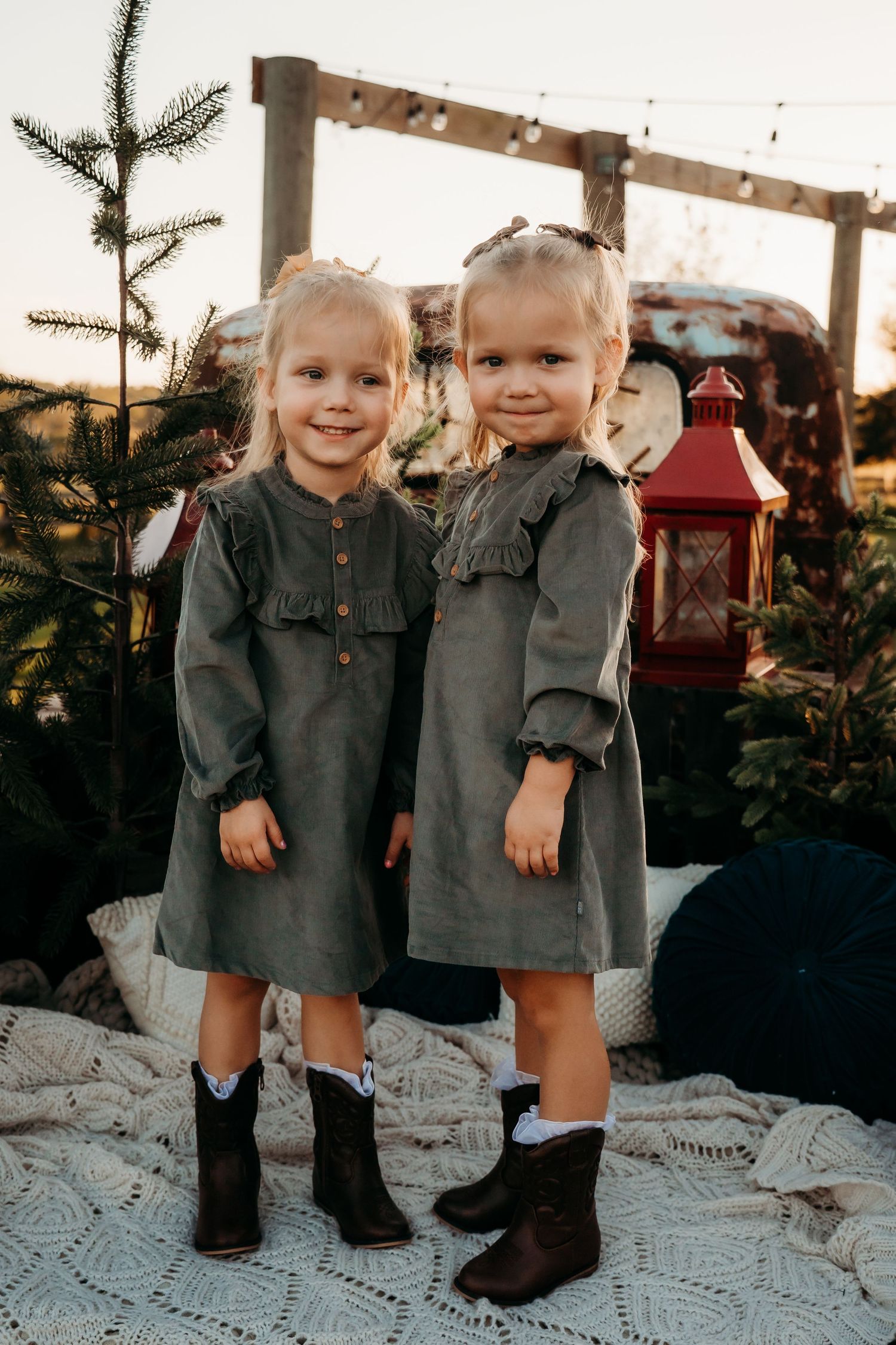 Adorable twin toddler girls in matching gray dresses and cowboy boots posing at golden hour outdoors.