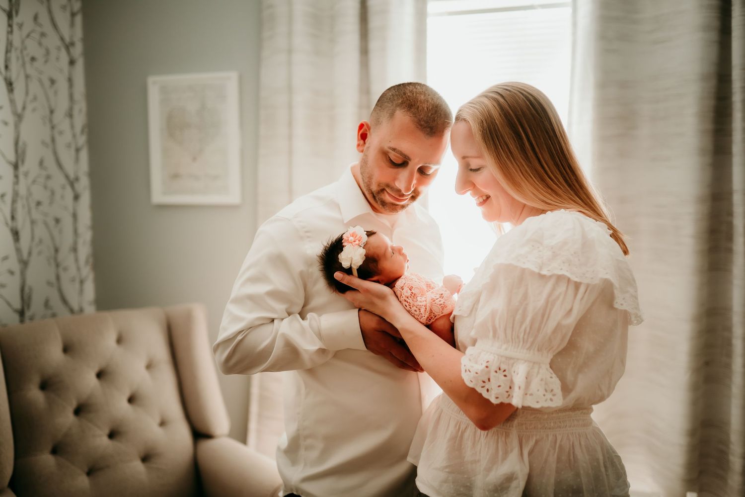 New parents in white outfits smiling at their newborn baby in a bright airy nursery.