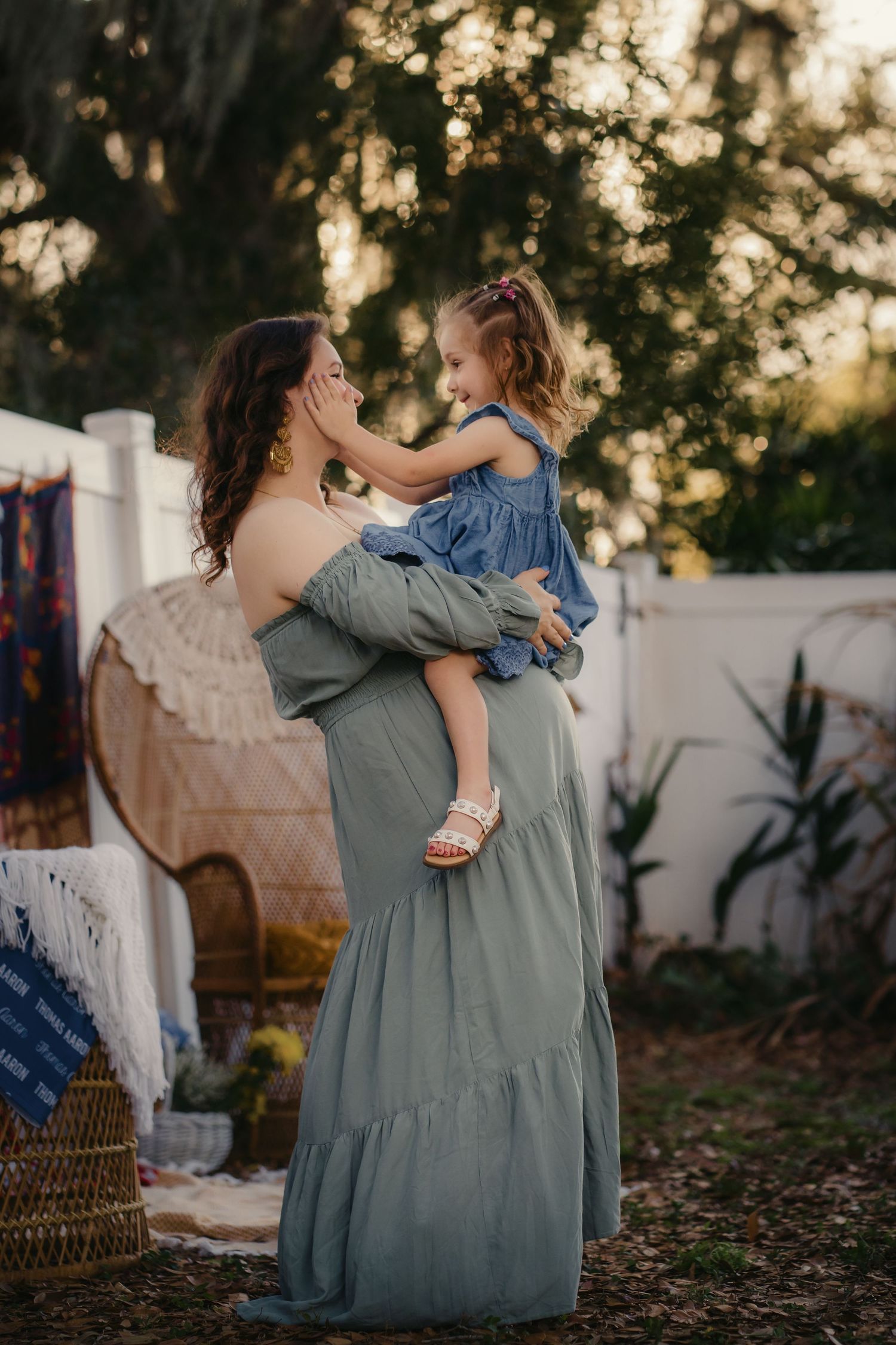 A mother in a sage green dress lifts her young daughter in a blue outfit outdoors at golden hour.