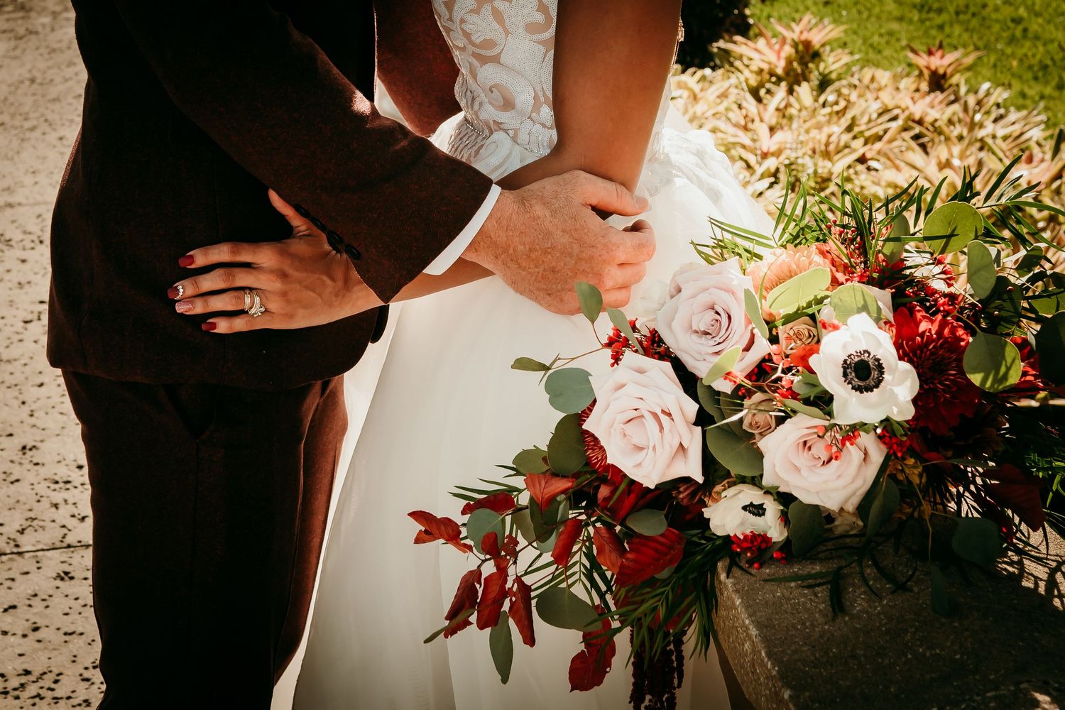 Bride and groom embracing while holding a lush bouquet of pink and red flowers with greenery at their wedding.