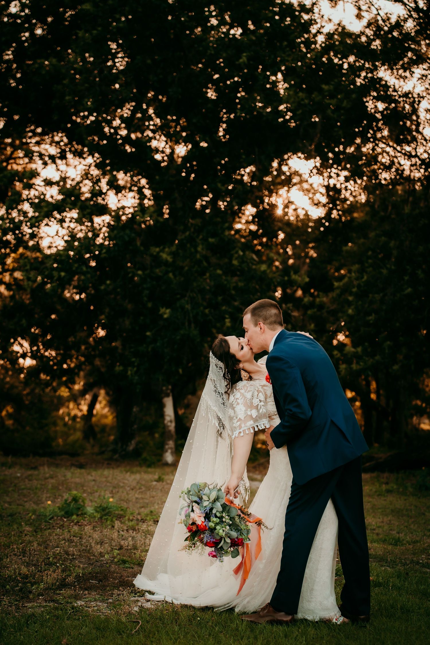 Bride and groom share a romantic kiss at golden hour sunset, surrounded by lush trees, as bride holds colorful bouquet.