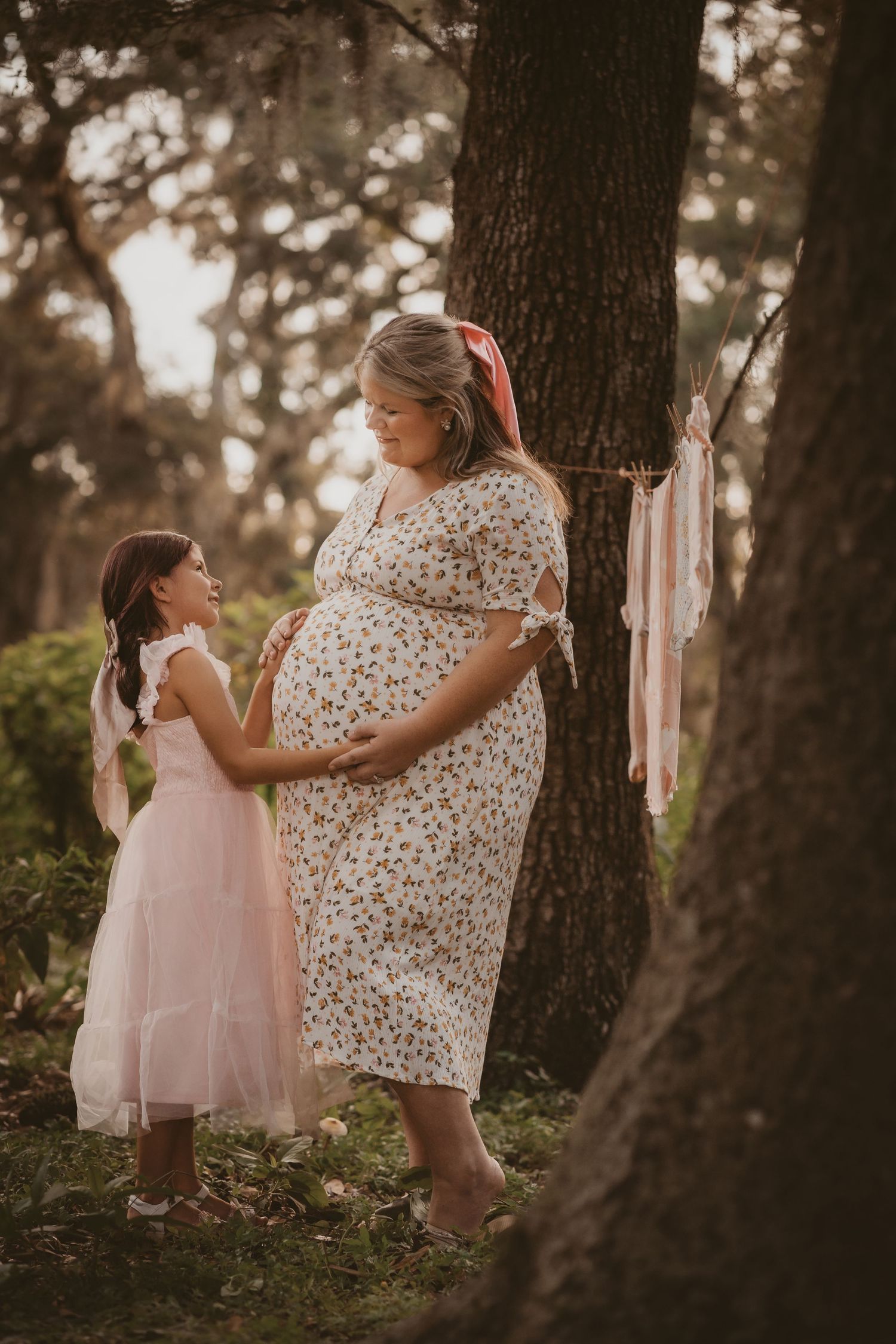 Pregnant woman in floral dress shares sweet moment with young girl by tree at sunset.