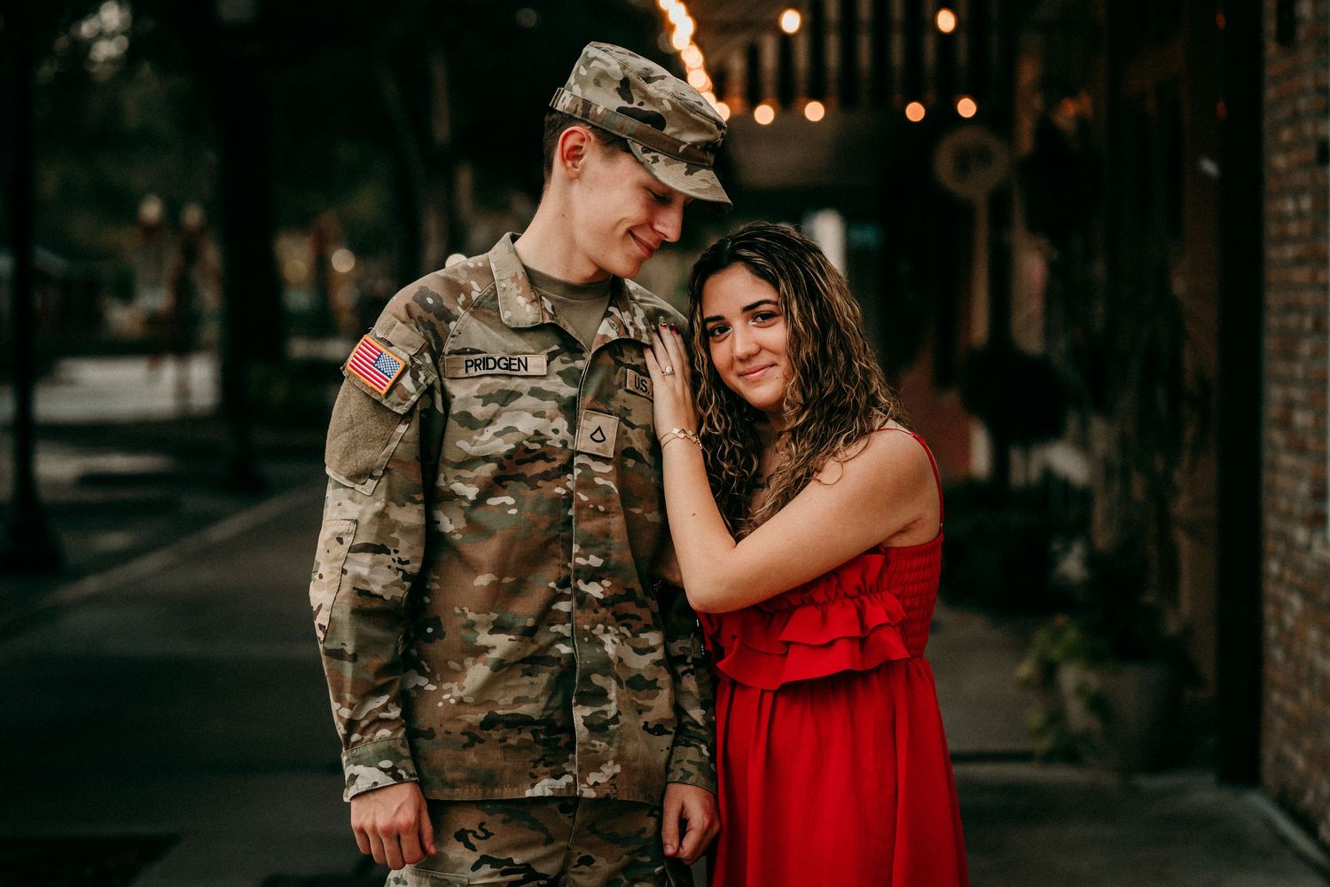 US Army soldier in camouflage uniform standing with woman in red dress on a city street.
