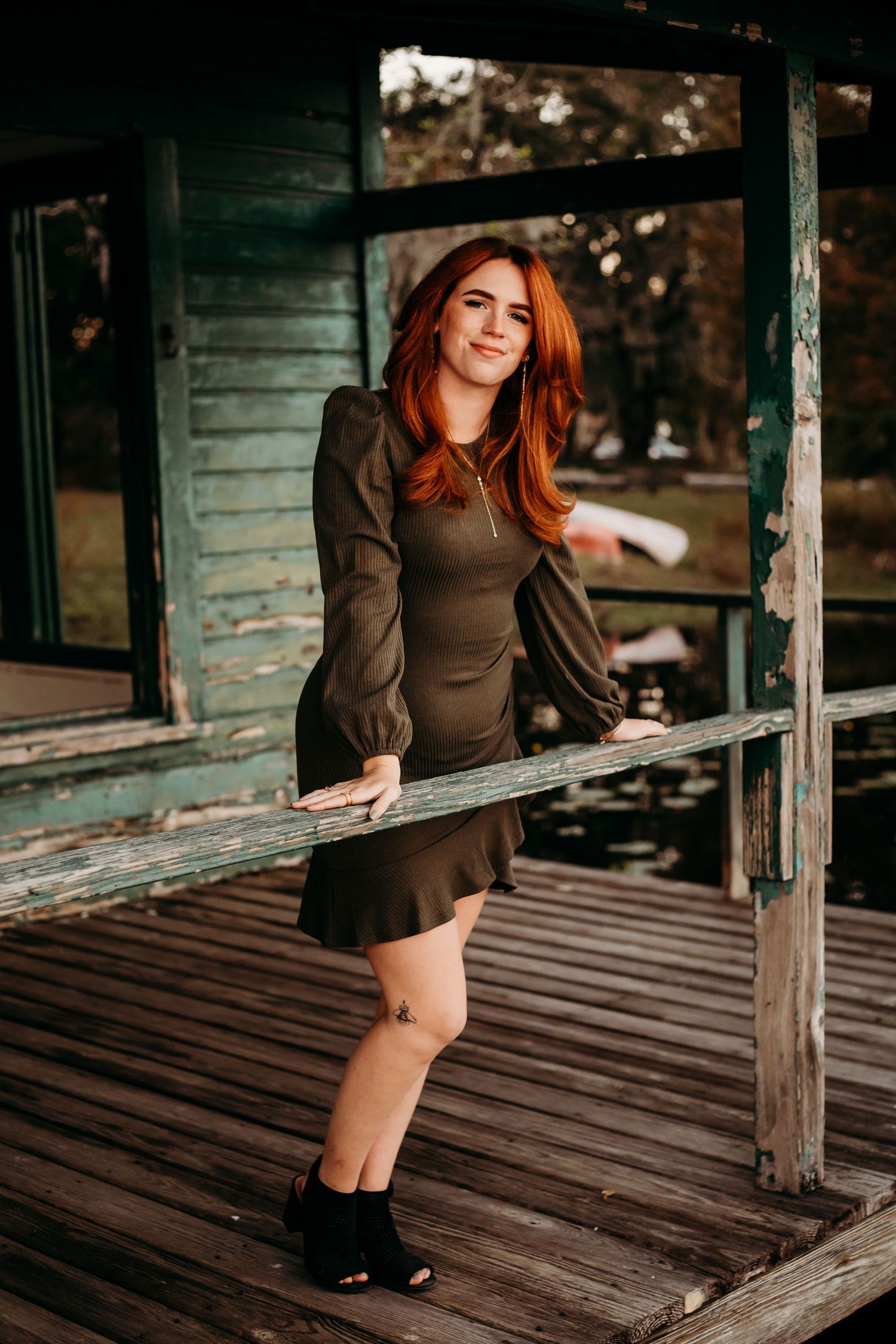 A young redhead woman in a dark mini dress and black heels poses on a rustic wooden porch.