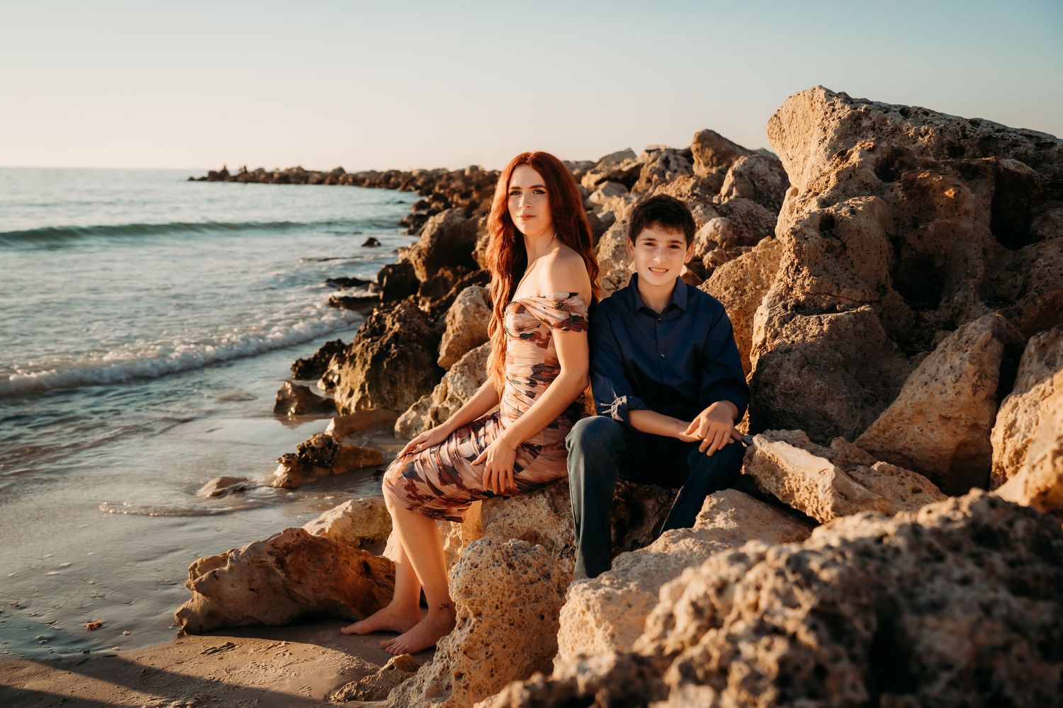 Redhead woman and young boy sitting on coastal rocks at sunset.