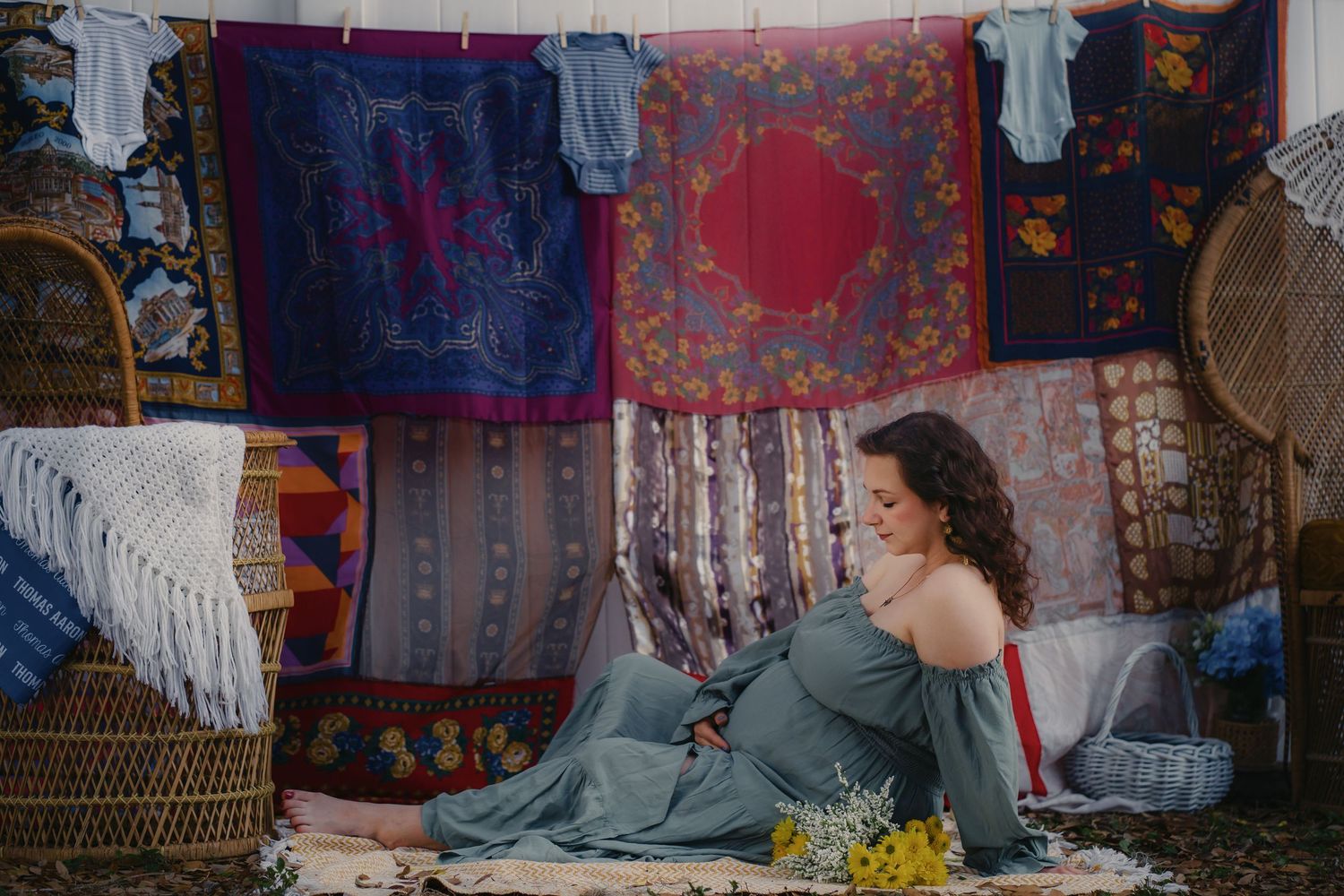 Pregnant woman in blue dress sits among colorful bohemian tapestries and wicker baskets with yellow flowers. Lifestyle family photographer