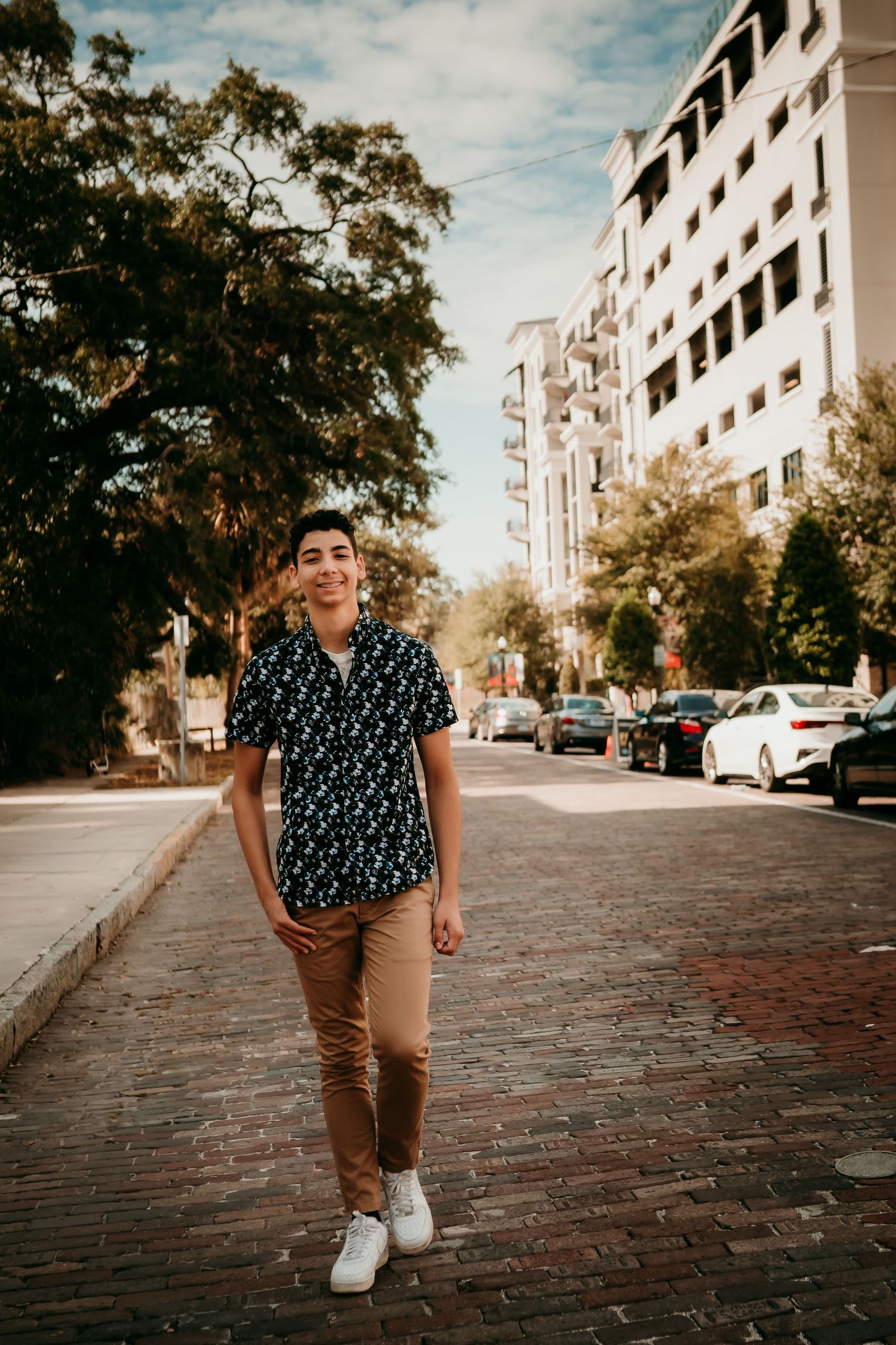 Young man in patterned shirt and tan pants walks on brick sidewalk in urban setting with trees and buildings.