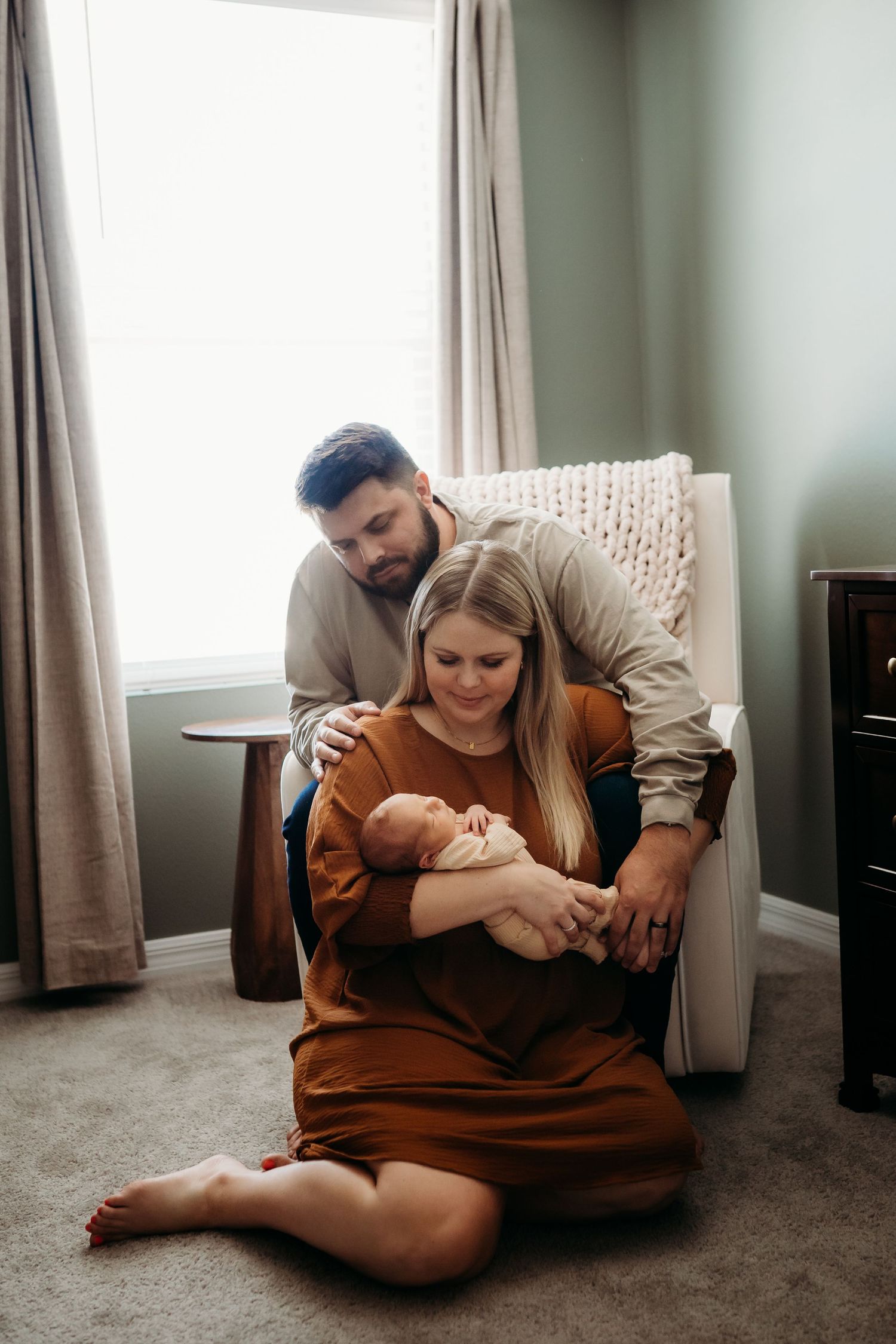 New parents sitting on nursery floor together, mother cradling sleeping newborn in her arms.