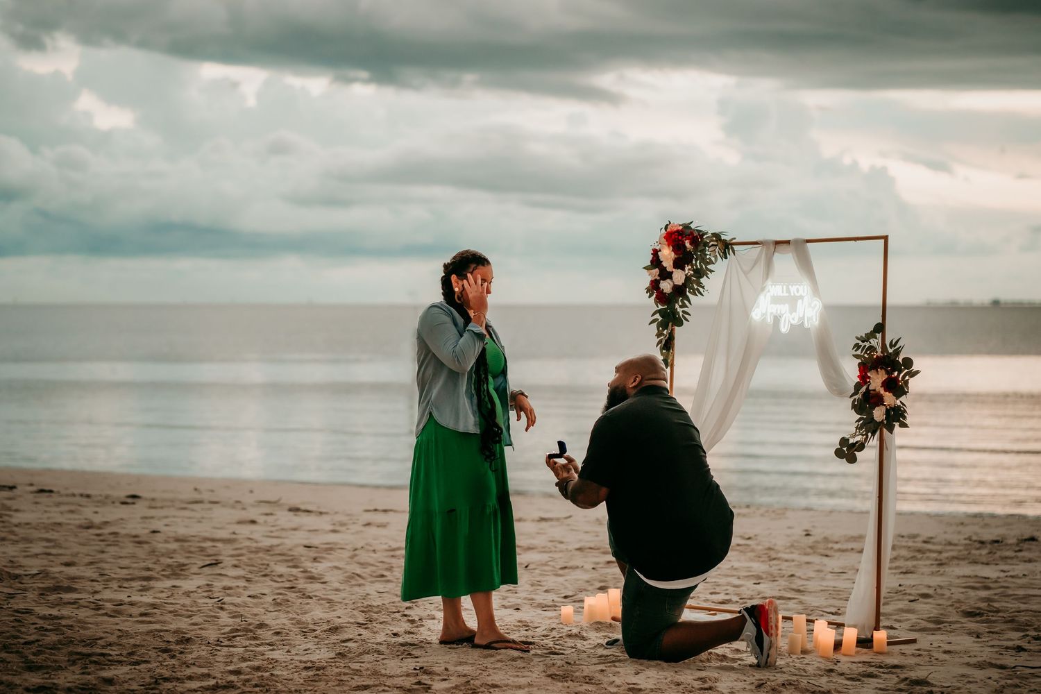 Man proposes to woman on beach at sunset near floral arch with candles, romantic seaside marriage proposal.