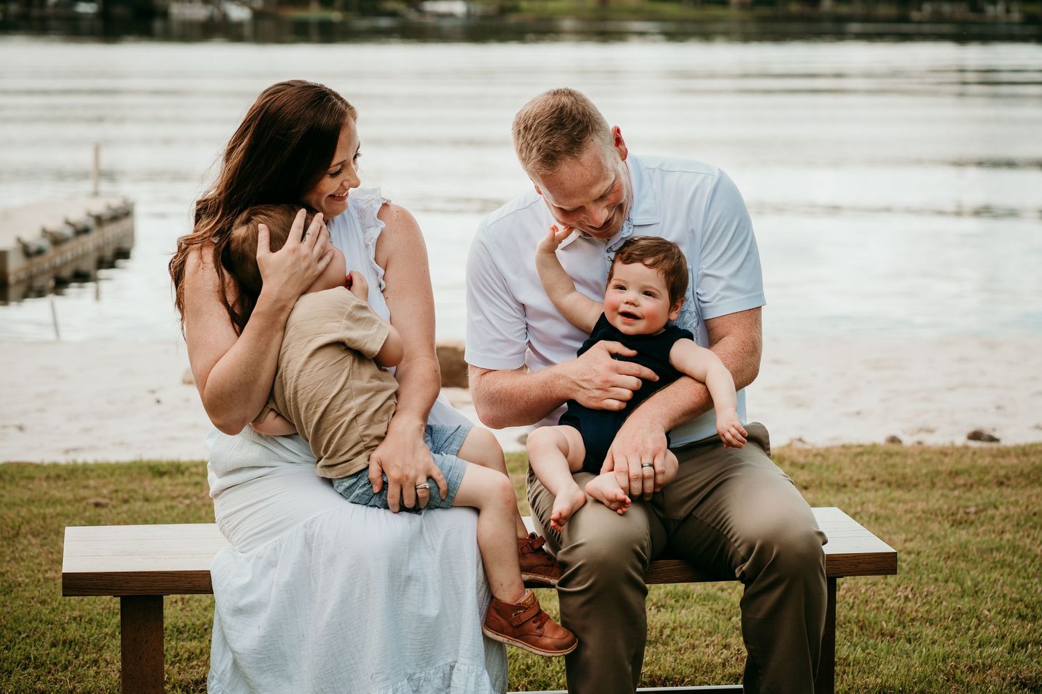 Happy family of four sitting on a bench by a lake, parents smiling at their two young children.