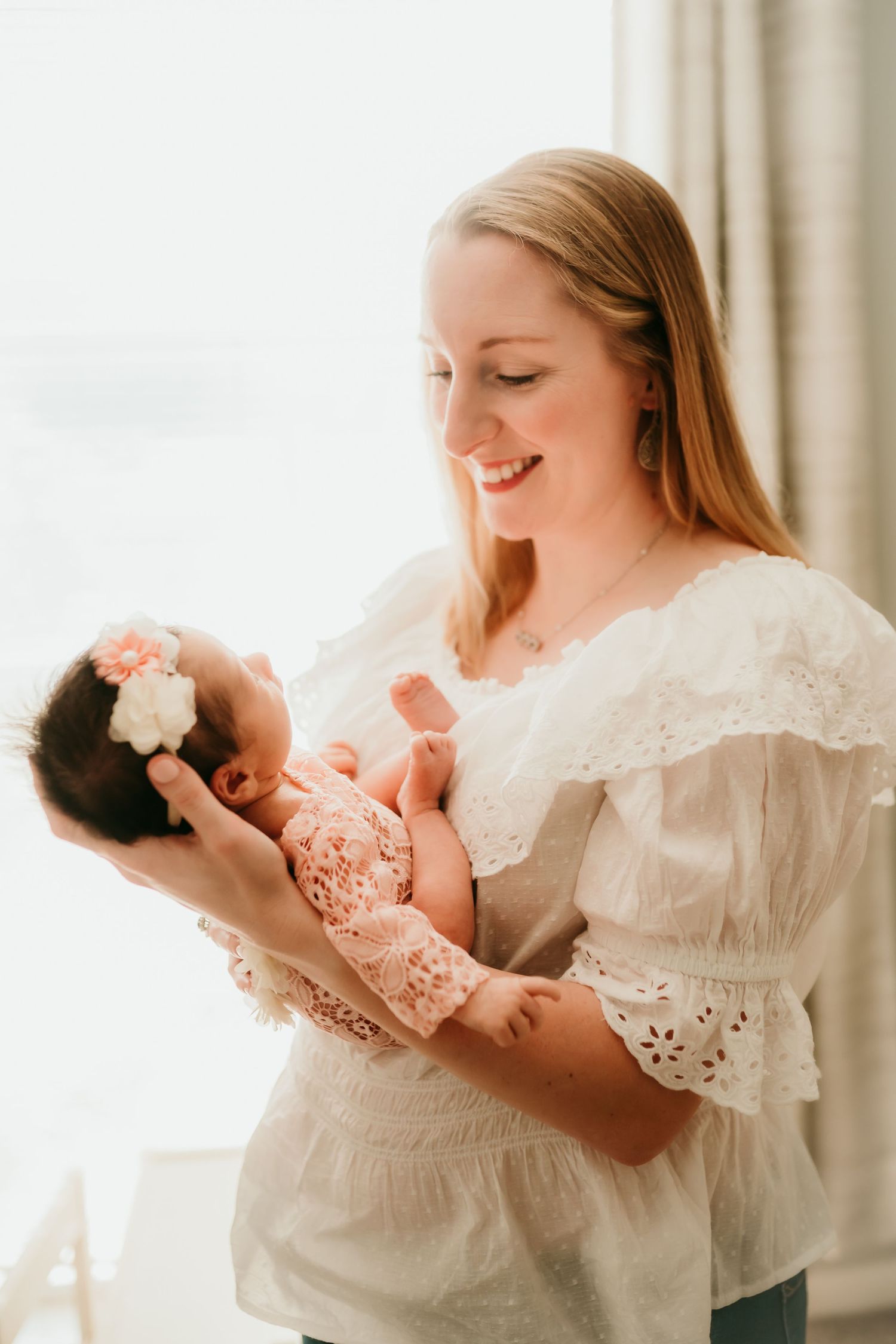 Smiling blonde mother in white lace top cradling her newborn baby girl with floral headband.