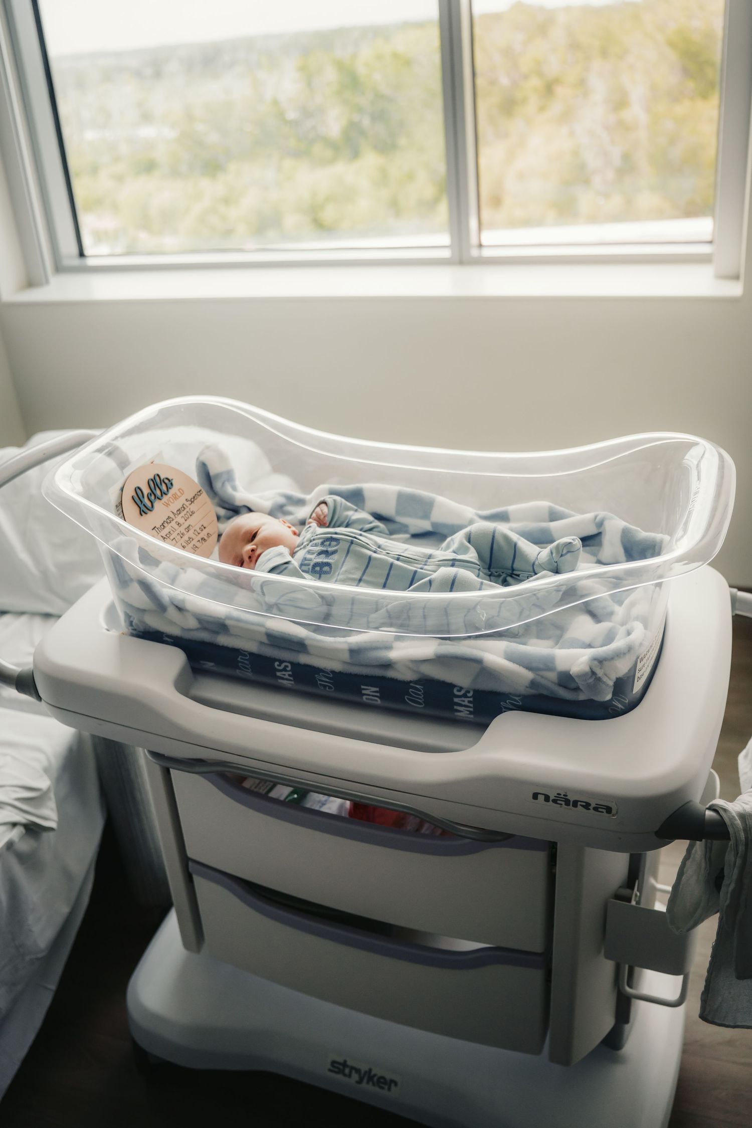 Newborn baby resting in a hospital bassinet on a Stryker cart near a window.