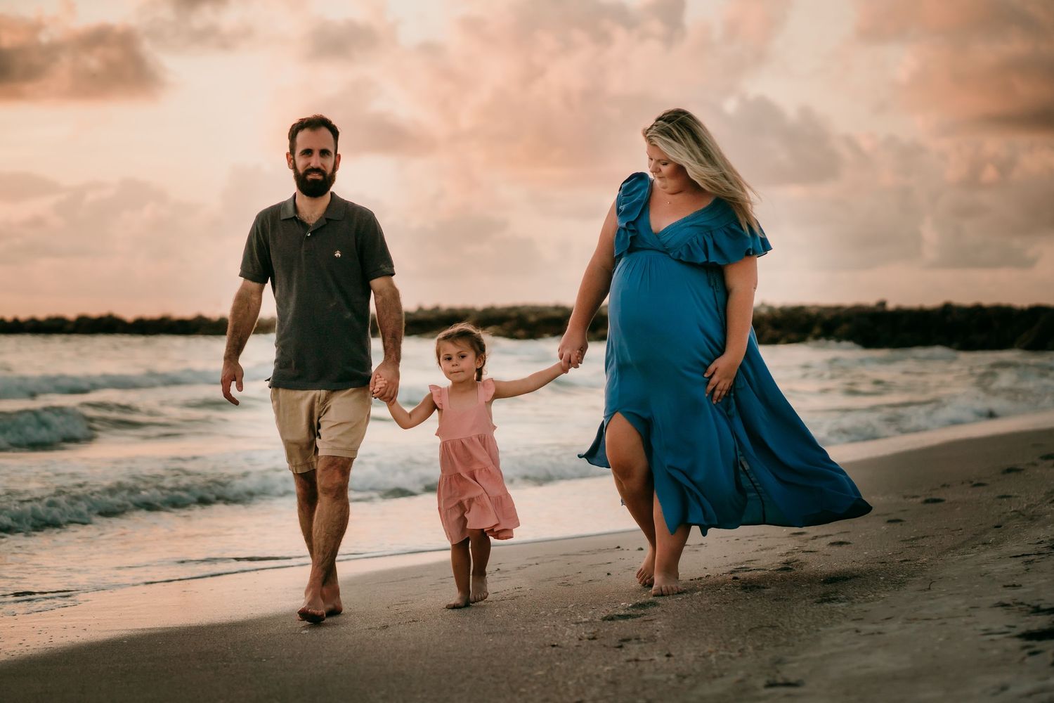 Pregnant woman in blue dress walks on beach at sunset with bearded husband and young daughter.