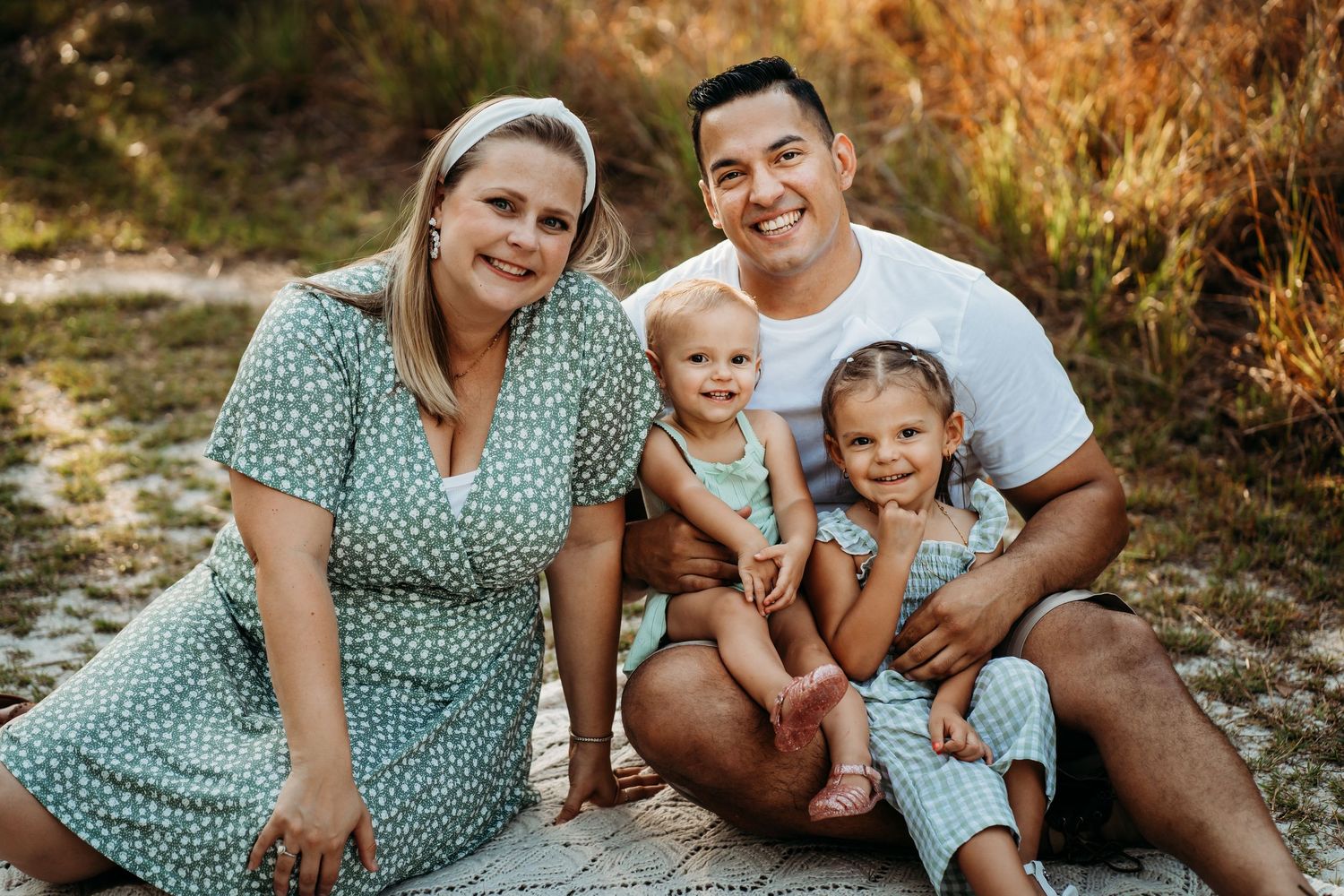 Happy family of four in matching green outfits sitting together on sandy ground outdoors.