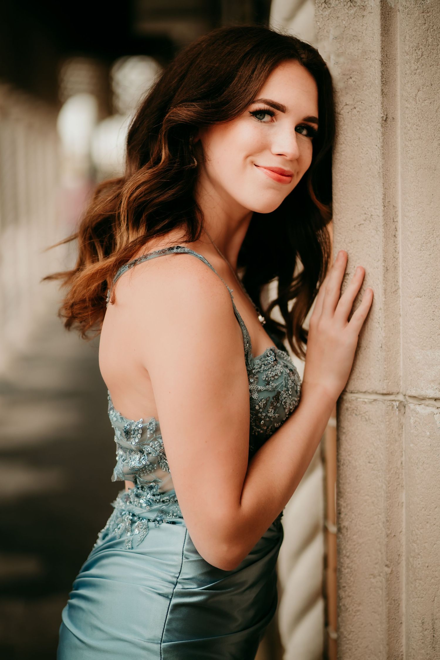 Young woman in elegant teal sequined gown leaning against stone column, smiling gracefully for a portrait photo.