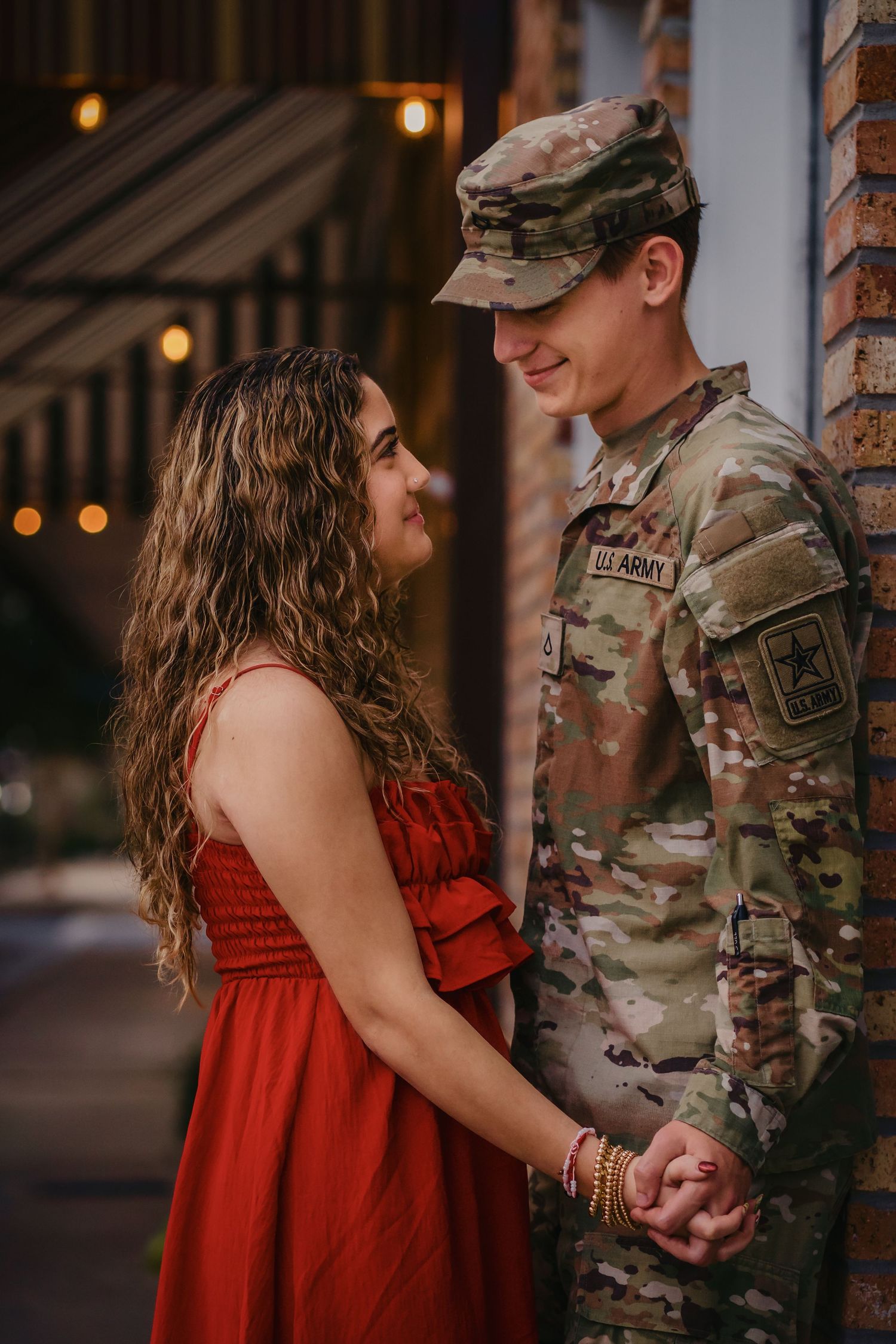 A couple holds hands tenderly, the man in a U.S. Army uniform and the woman in a red dress, sharing a romantic moment.