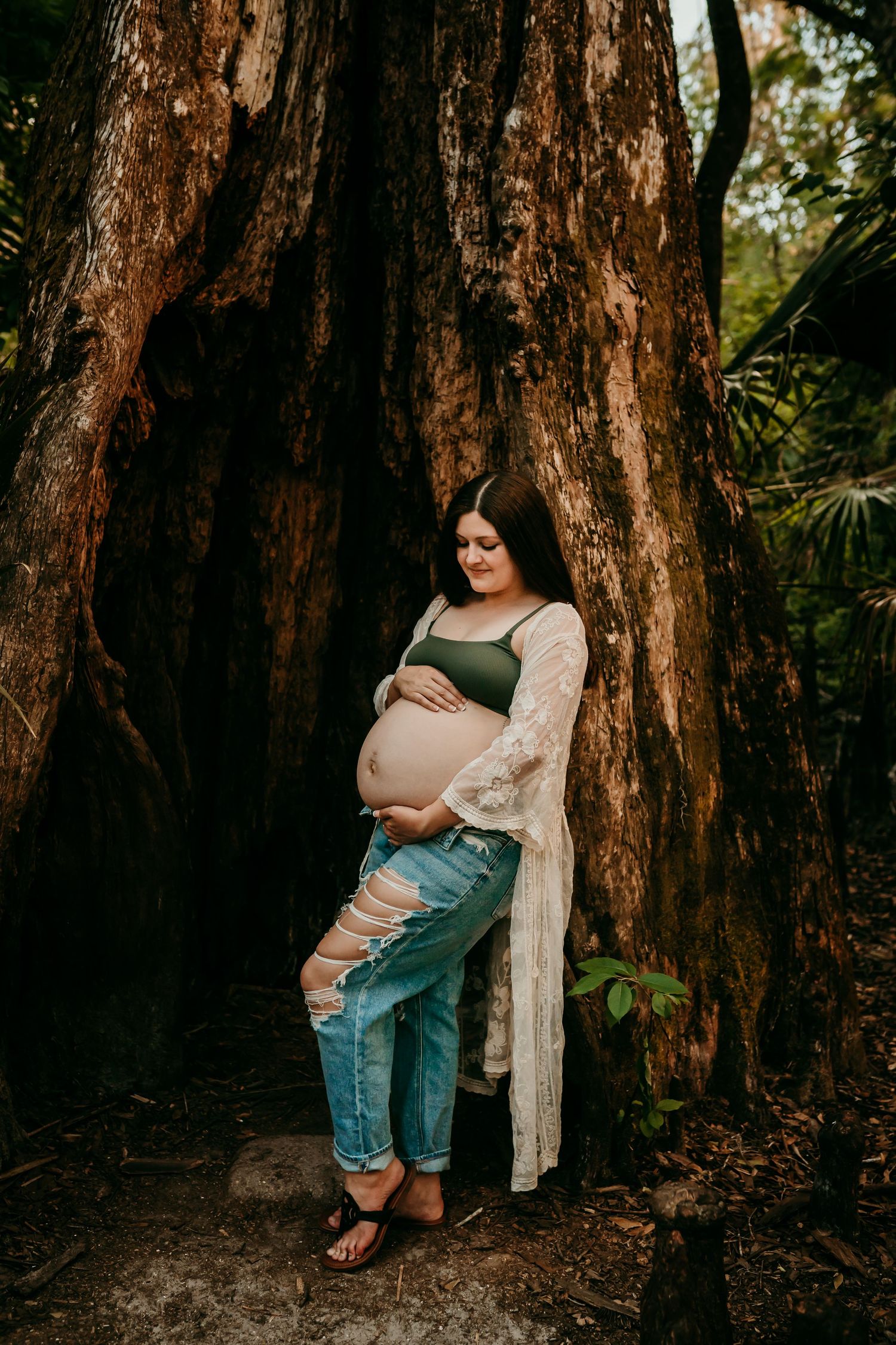 Pregnant woman in green bralette and ripped jeans leaning against a large tree outdoors.
