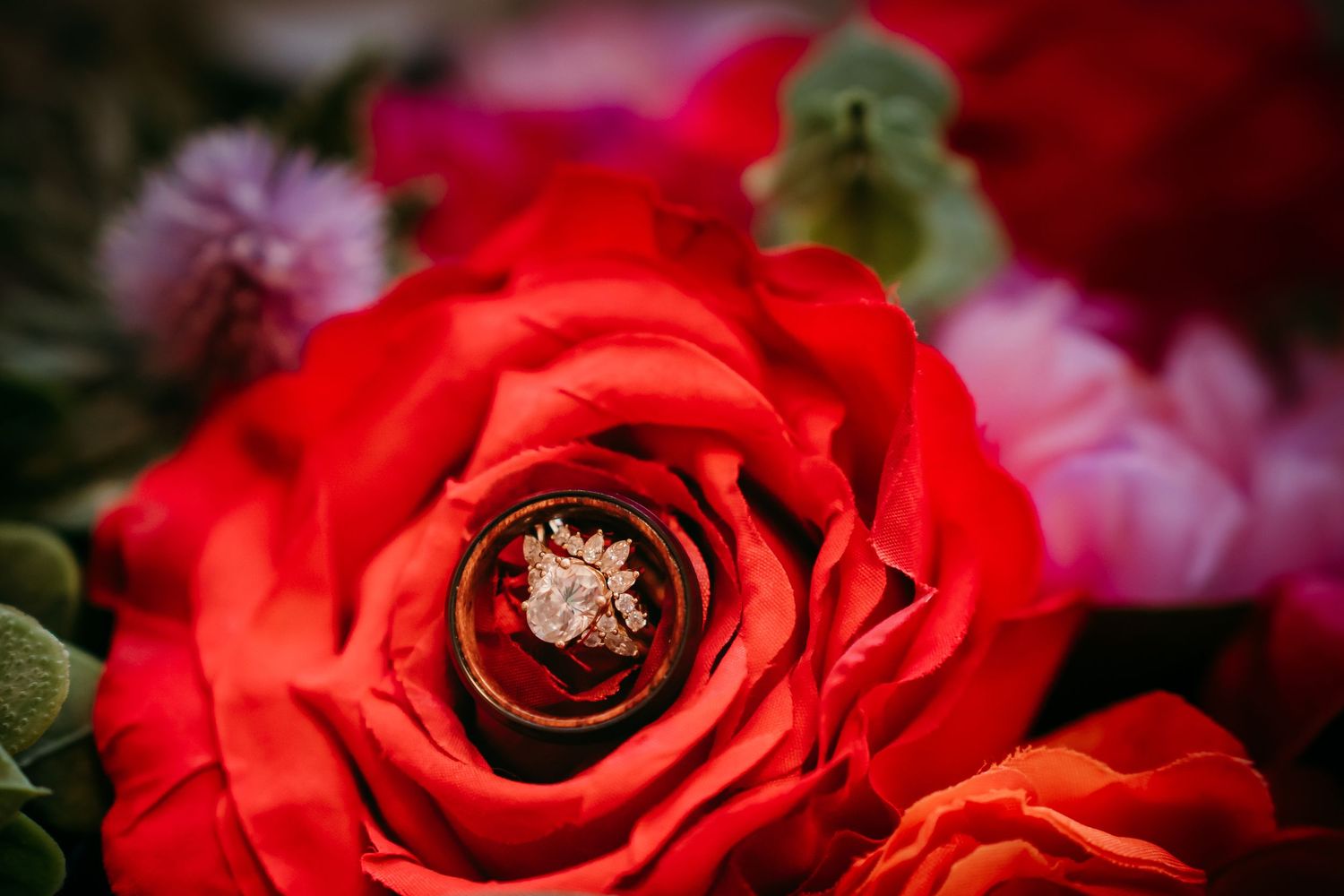 Gold wedding rings nestled inside a vibrant red rose bloom surrounded by colorful flowers in a bridal bouquet.