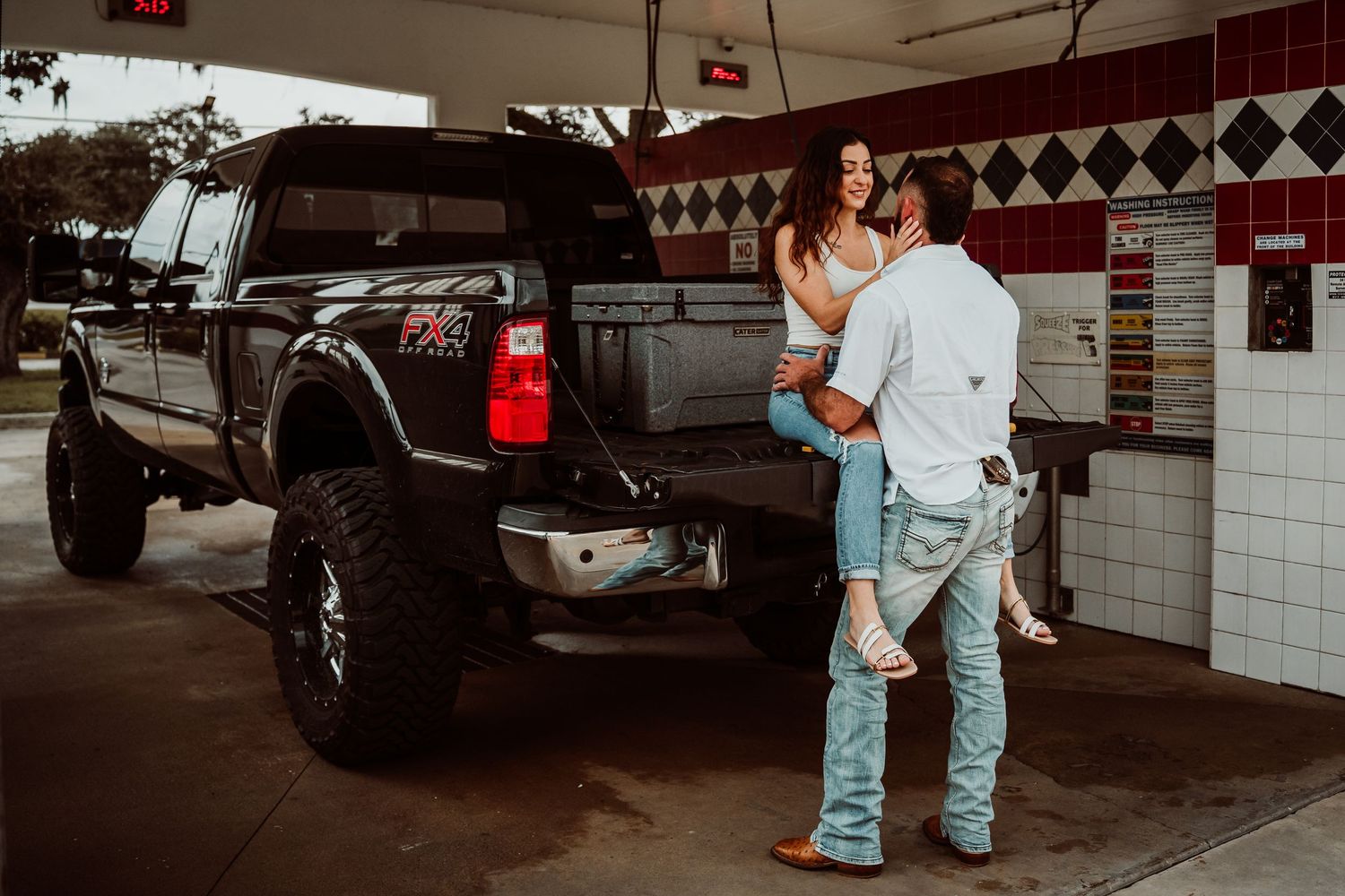 Couple embracing near lifted black Ford truck at outdoor gas station in casual jeans.