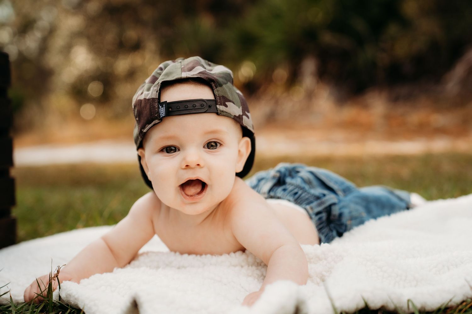 Adorable baby in camo snapback hat and jeans lying on white fur blanket outdoors, smiling at camera.