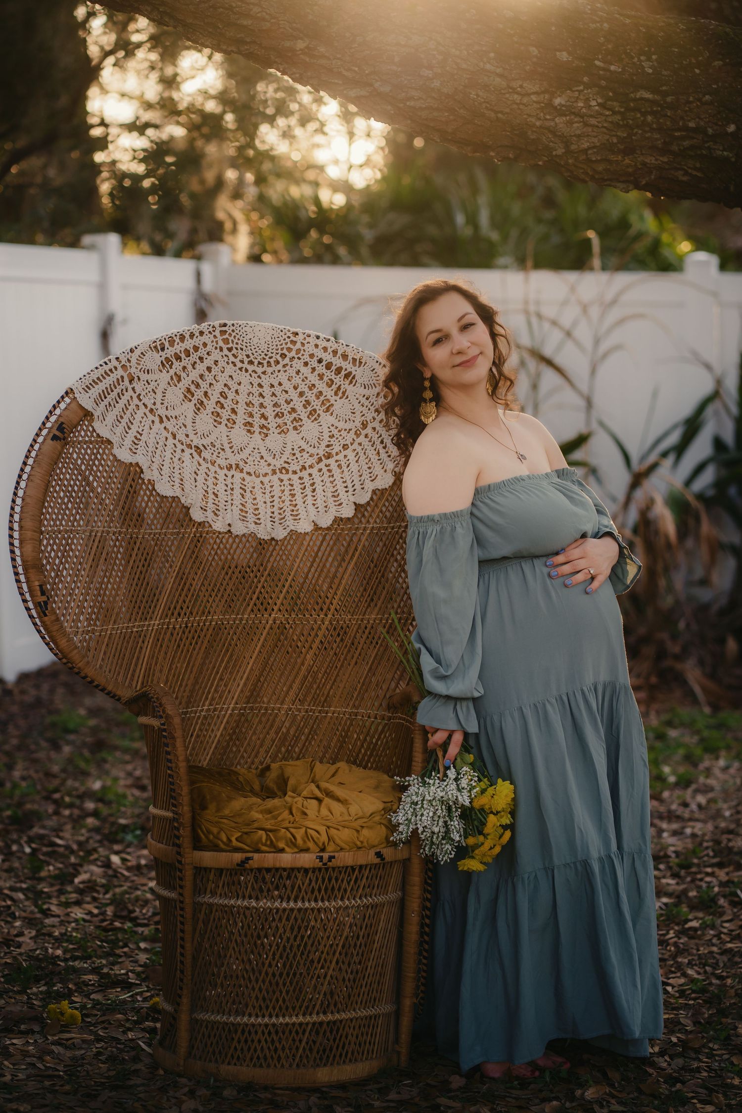 Pregnant woman in sage green off-shoulder gown holding flowers beside wicker peacock chair at golden hour.