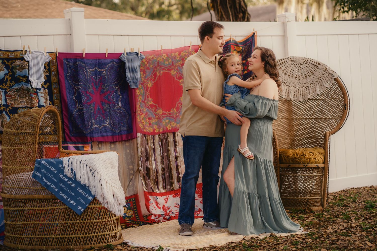 Family maternity photo shoot with colorful tapestry backdrop, wicker furniture, and boho outdoor setting.