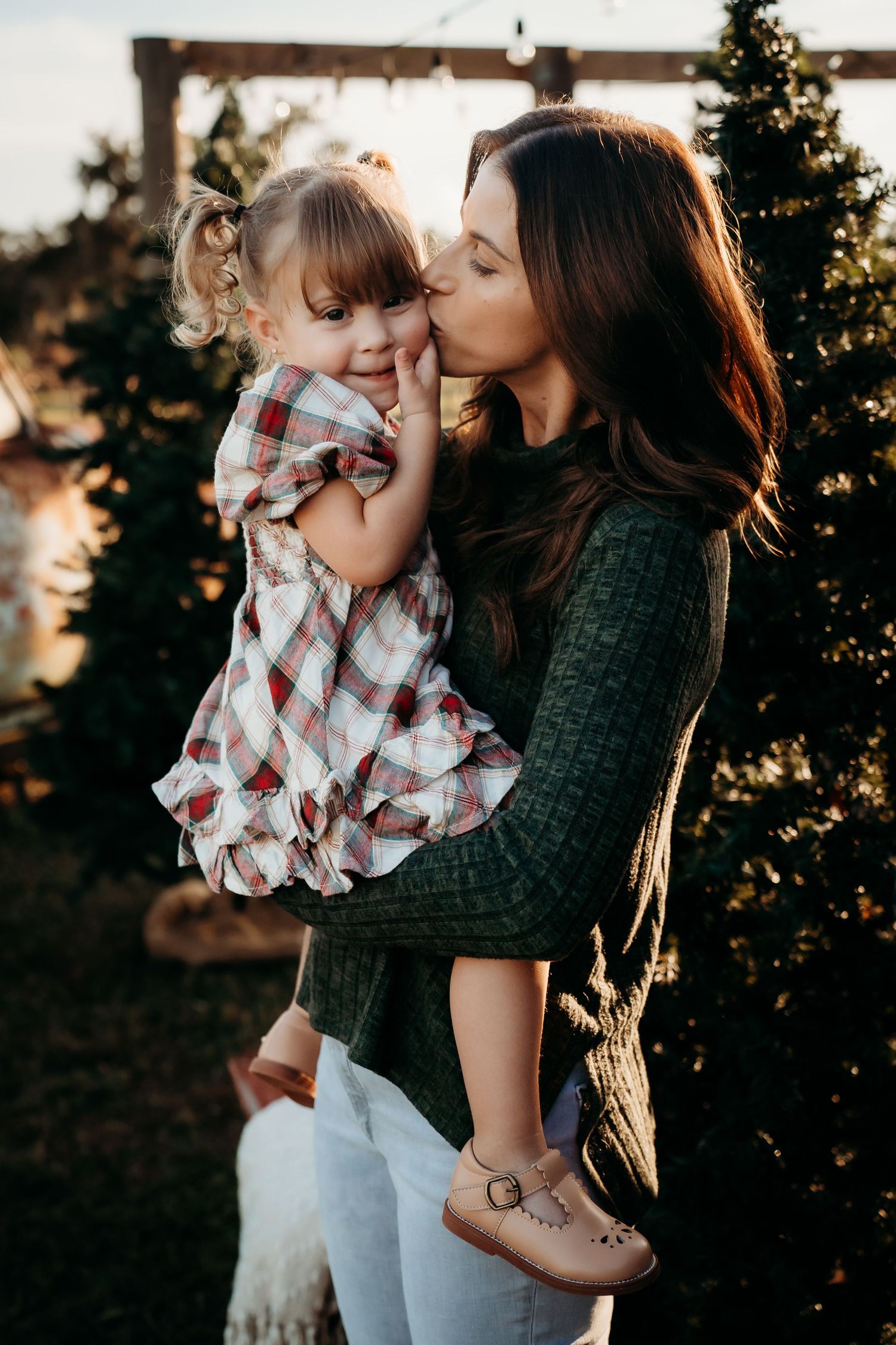 Mother kissing her toddler daughter on the cheek at a Christmas tree farm during golden hour.