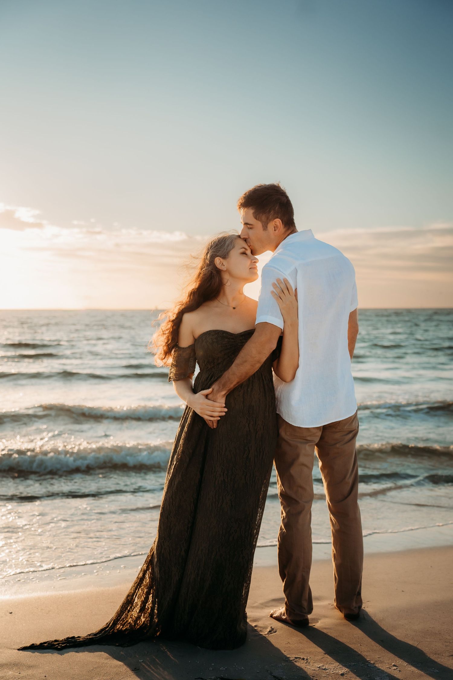 A couple shares a tender embrace on a sandy beach at golden hour, the woman wearing an elegant black maternity gown.